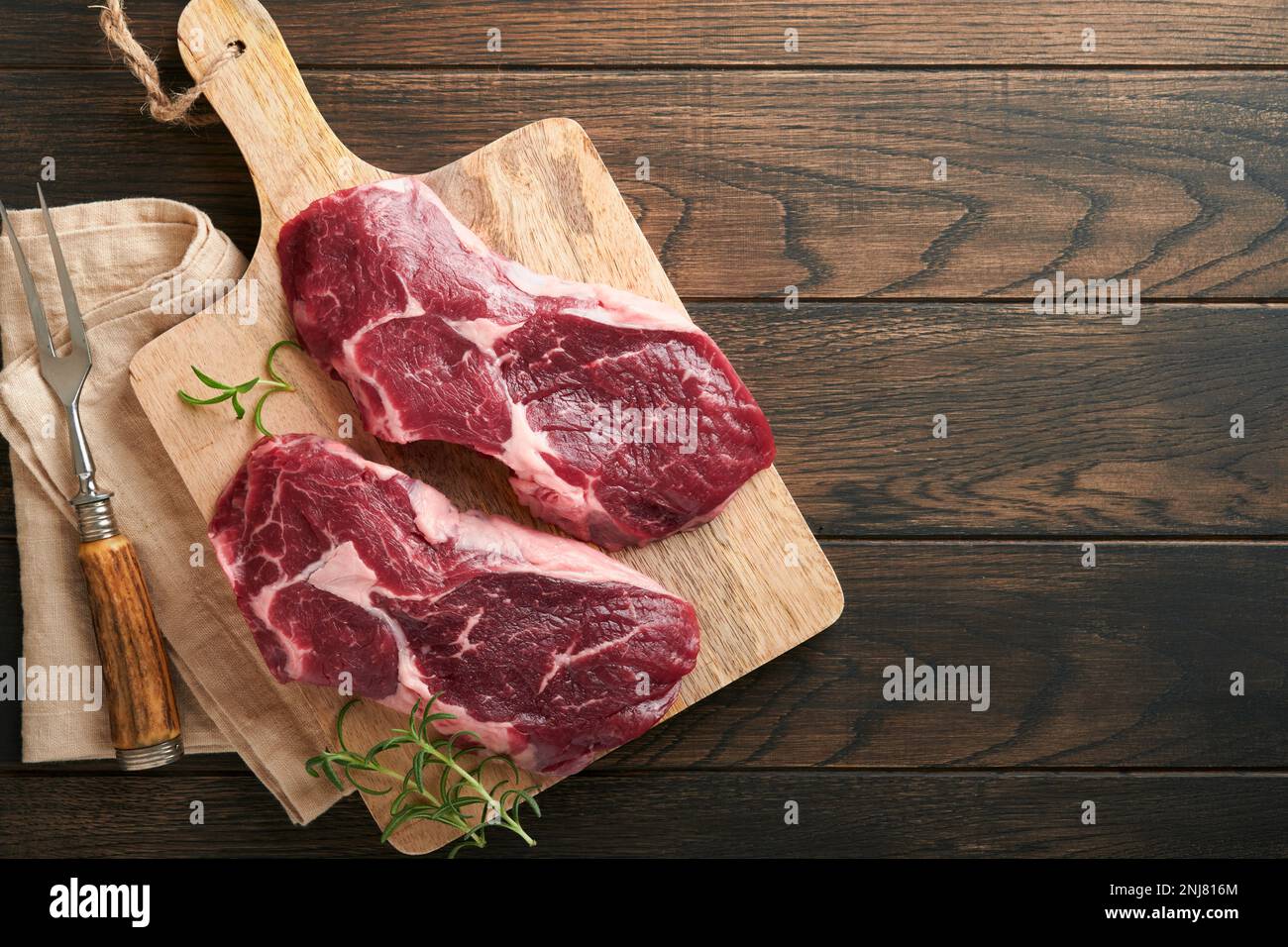 Rohes Rindersteak. Marmoriertes rohes Ribeye-Steak mit Rosmarin, Salz und Pfeffer auf dem Schneidebrett auf dunklem, altem Holzhintergrund. Rohes Rindersteak und Gewürze Stockfoto