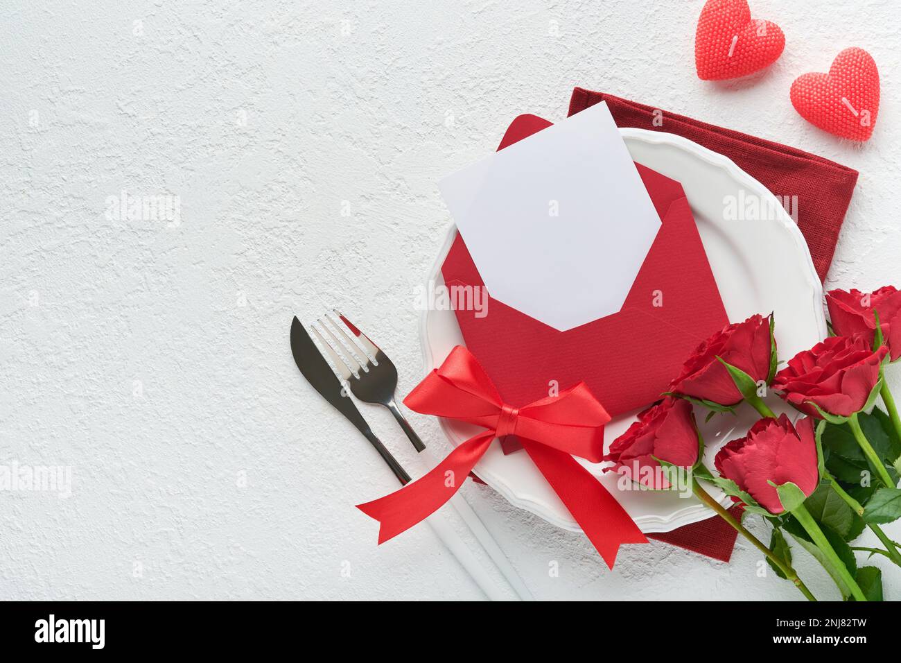 Romantische kreative Tischgestaltung. Leerer weißer Teller und rote Rosen, Kerzen, Messer, Gabel und dekorative Seidenherzen auf weißem Hintergrund. Hochzeit Zum Valentinstag Stockfoto