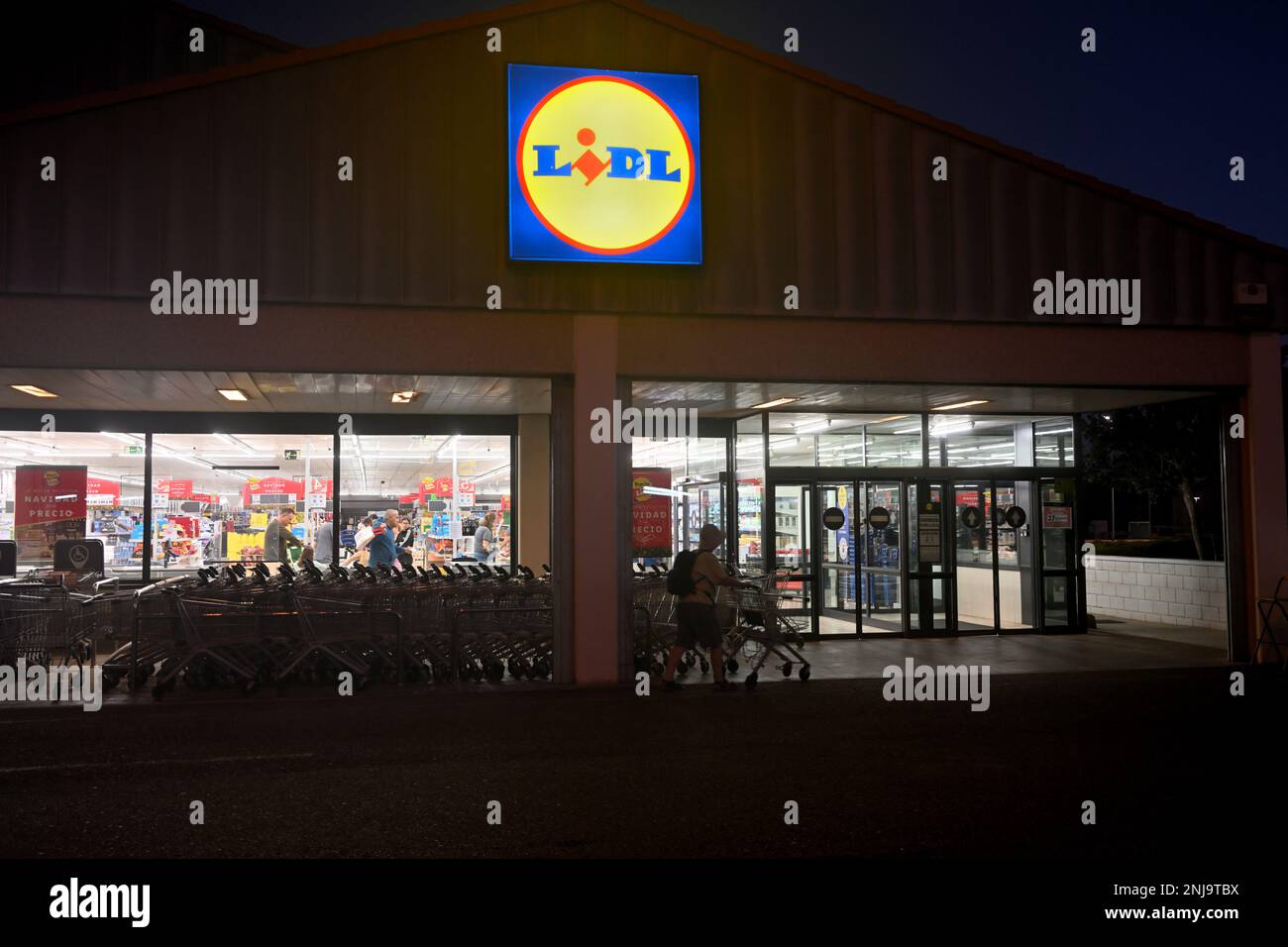 Vor einem Lidl-Einzelhandelsgeschäft bei Nacht mit beleuchtetem Innenraum und Silhouetten von Einkaufswagen und Kunden draußen Stockfoto