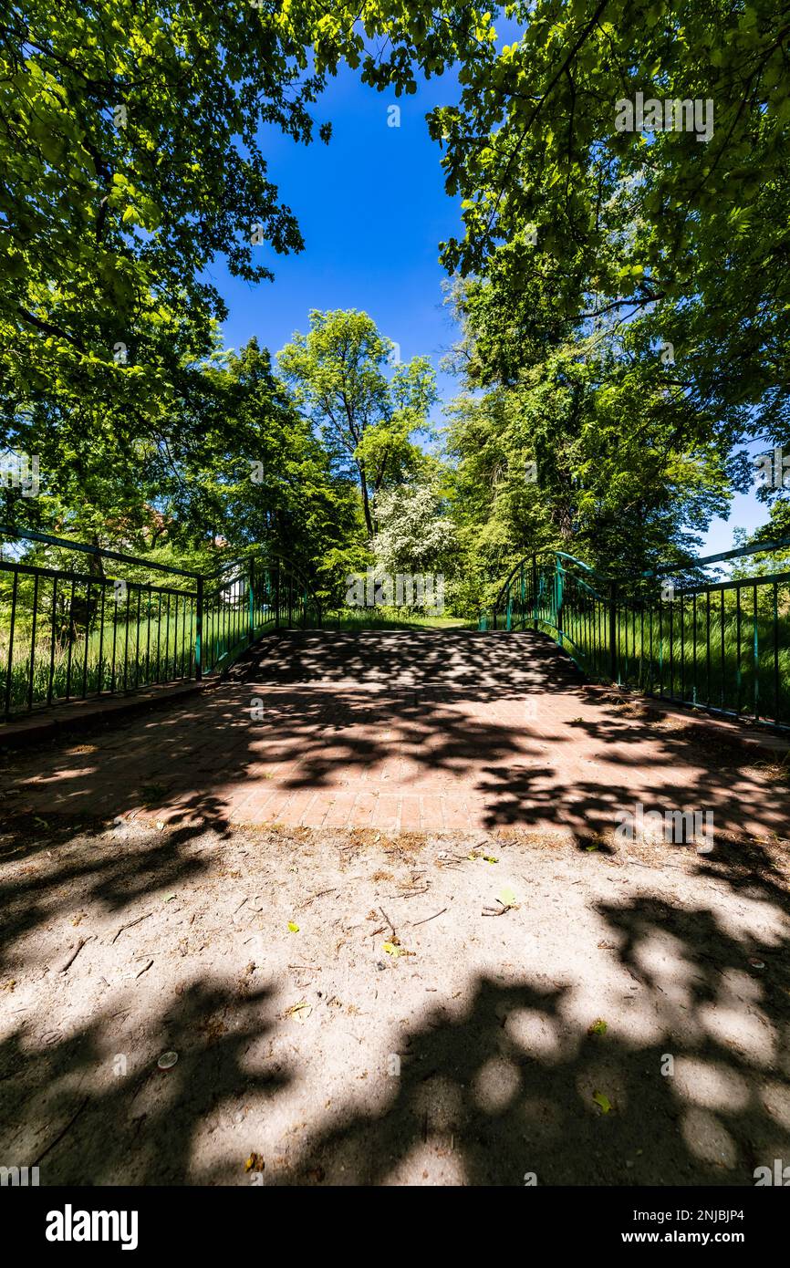 Alte renovierte Retro-Brücke mit grünen Metallgeländern über einen kleinen Fluss in einem kleinen bunten Park am sonnigen Morgen Stockfoto