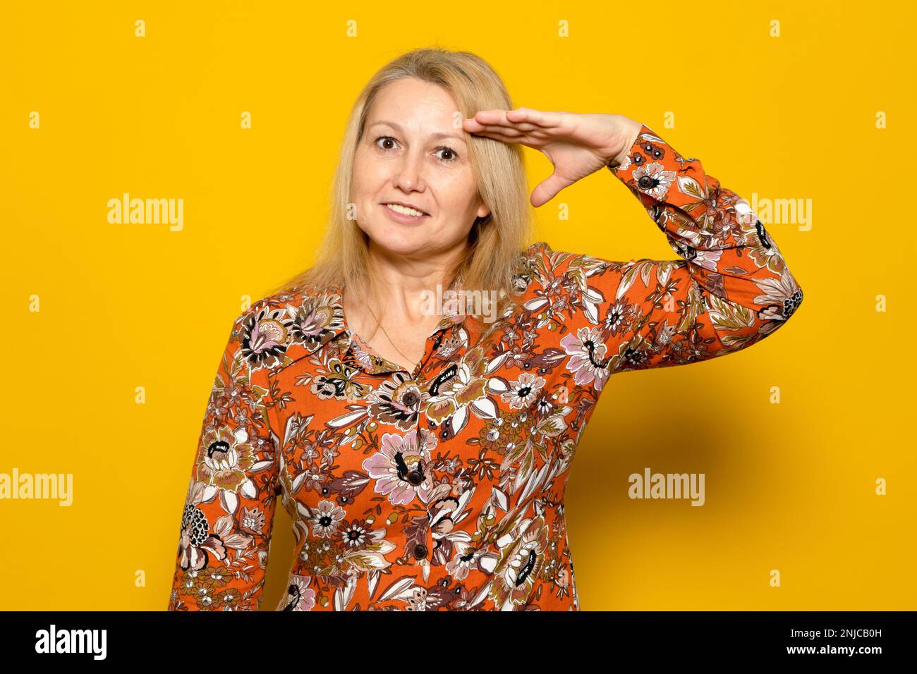 Weiße blonde Frau in einem gemusterten Kleid, die den Militärsalut abseits des gelben Hintergrunds macht, erfüllt die Heimat. Stockfoto