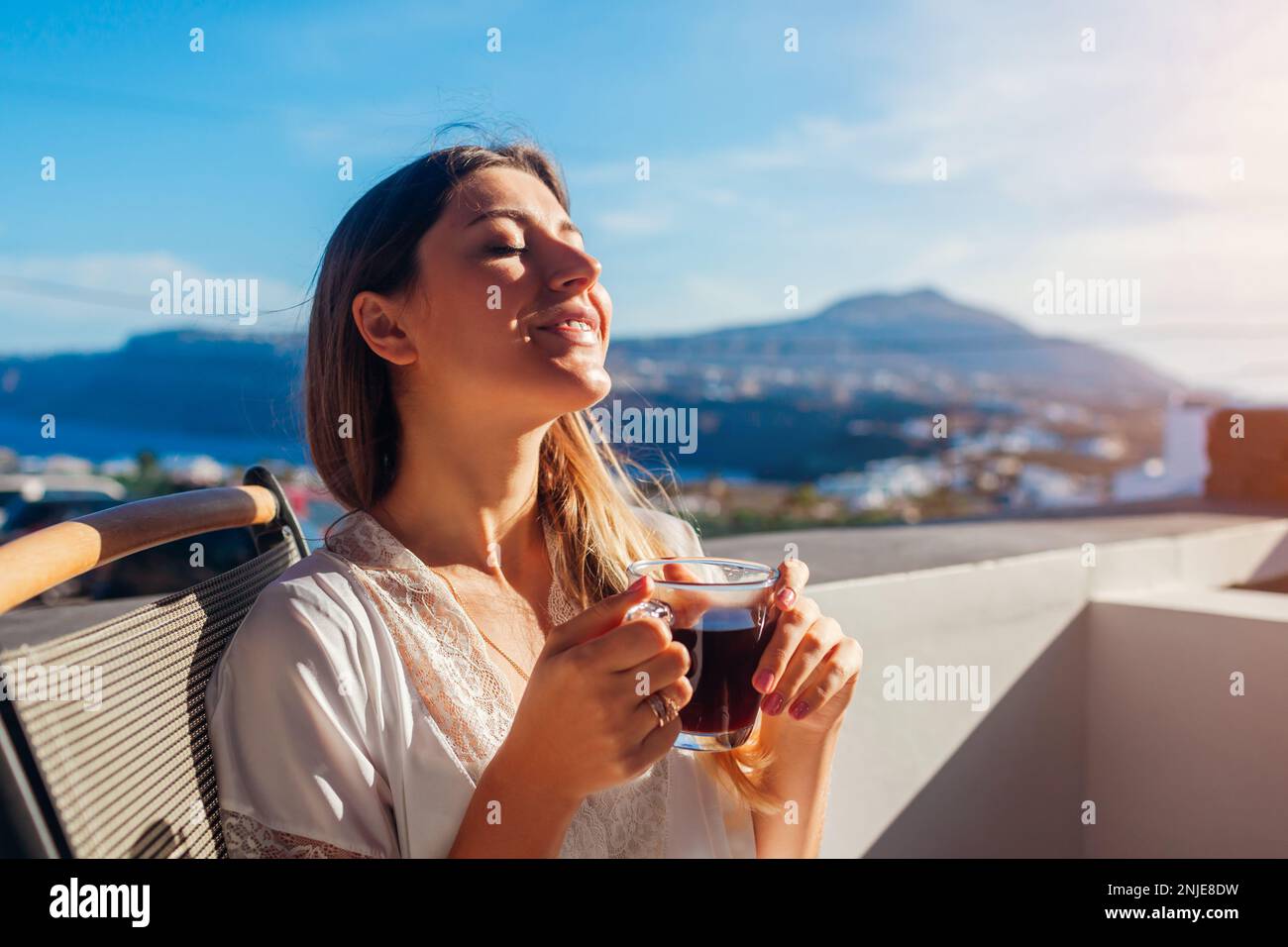 Eine Frau, die sich auf der Hotelterrasse entspannt und morgens Kaffee trinkt und die Berglandschaft von Santorin genießt. Frühstück mit Aussicht. Sommerferien Stockfoto Eine Frau, die sich auf der Hotelterrasse entspannt und morgens Kaffee trinkt und die Berglandschaft von Santorin genießt. Frühstück mit Aussicht. Sommerferien Stockfoto