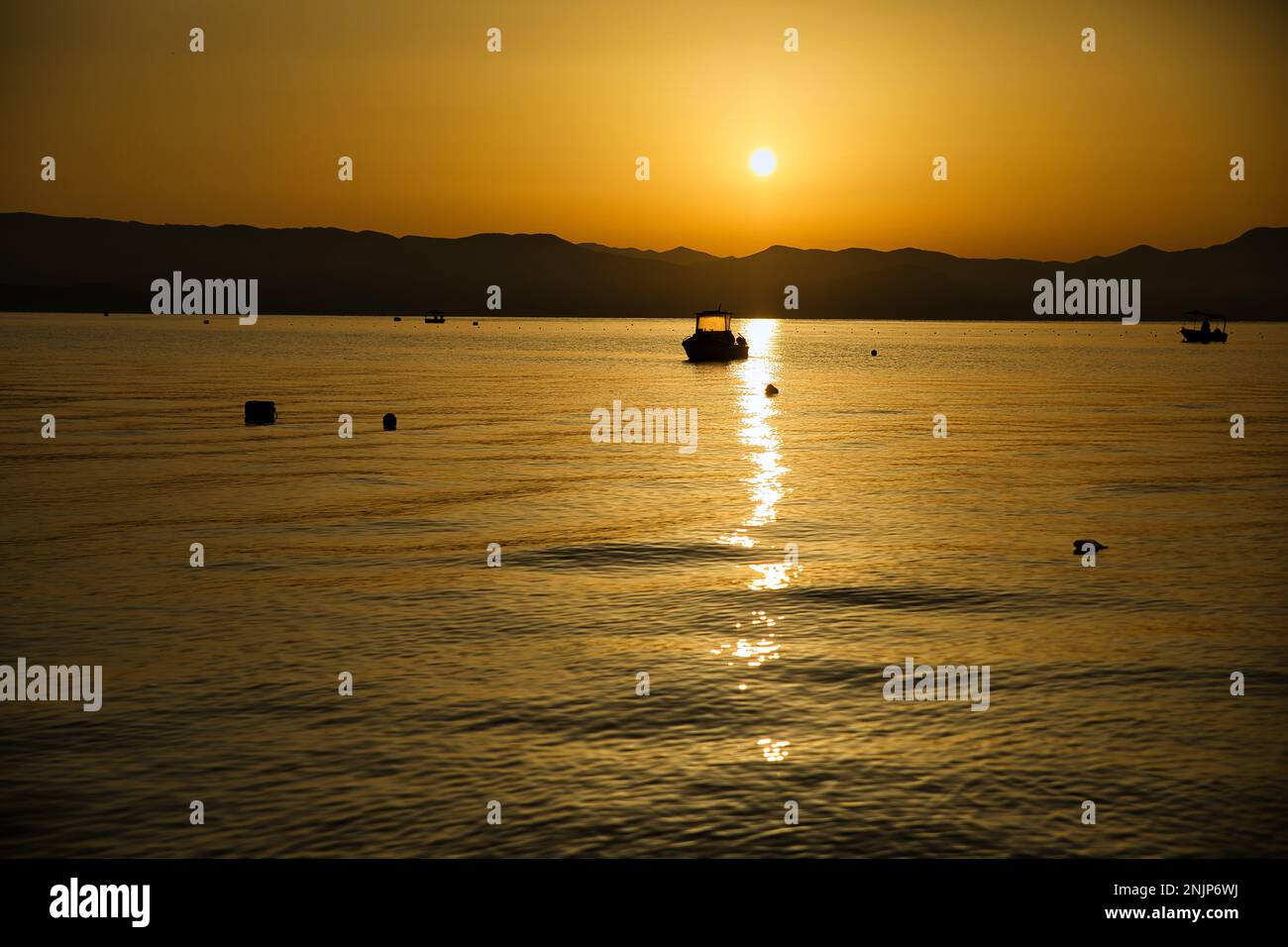 Sonnenuntergang über dem Meer auf einer Kette von Hügeln, der ganze Himmel scheint in goldgelben Farben, die Sonne spiegelt sich im Wasser, auf dem Meer ein kleines Boot. Stockfoto