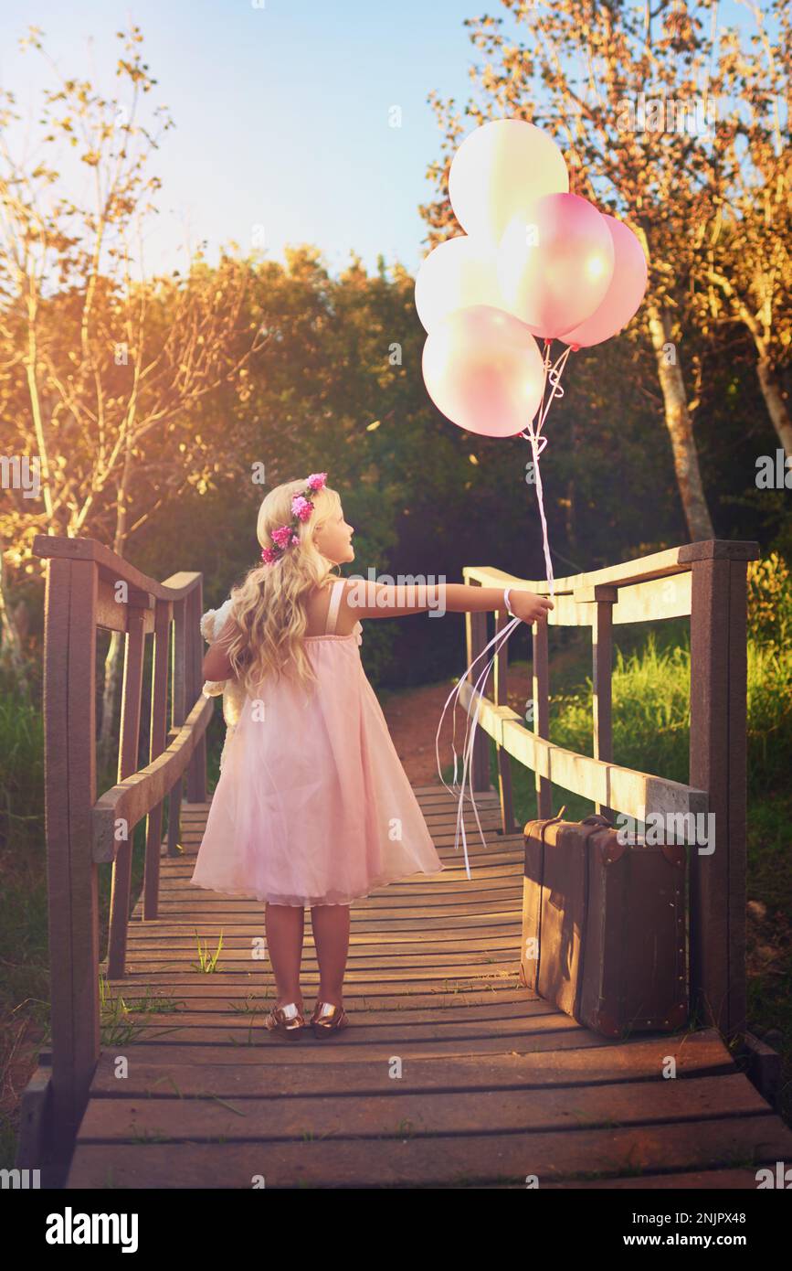Die Welt ist ihr Spielplatz. Ein glückliches kleines Mädchen, das Ballons und einen Teddybär hält, während es mitten auf einer Brücke steht. Stockfoto