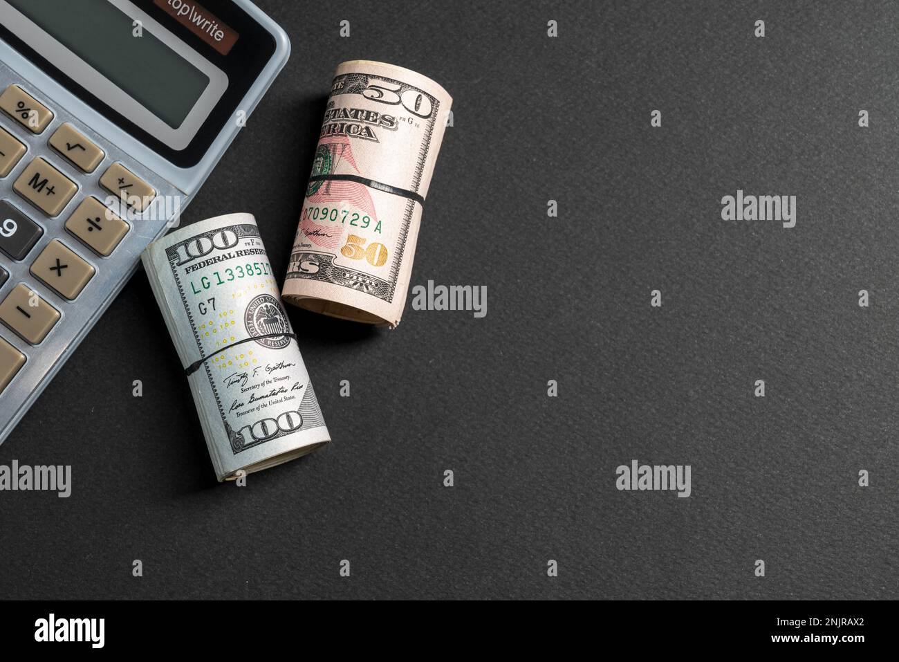 Rollen mit 50 und 100 US-Dollar stehen neben dem Rechner auf dunklem Hintergrund Stockfoto