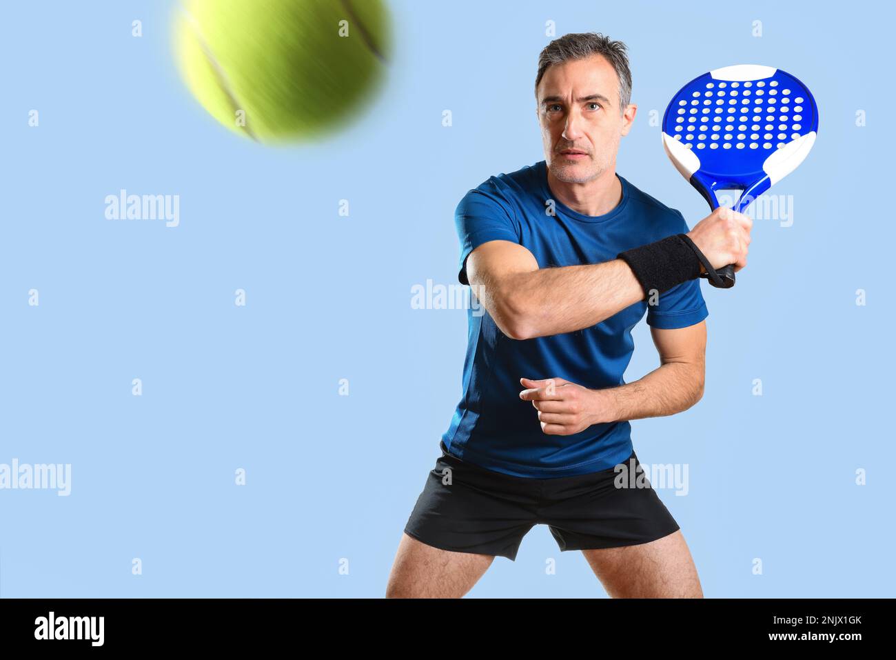 Porträt eines Mannes, der Paddeltennis spielt und einen Ball mit der Rückhand trifft. Er trägt blau-schwarze Sportkleidung und einen blau isolierten Hintergrund. Vorderansicht Stockfoto