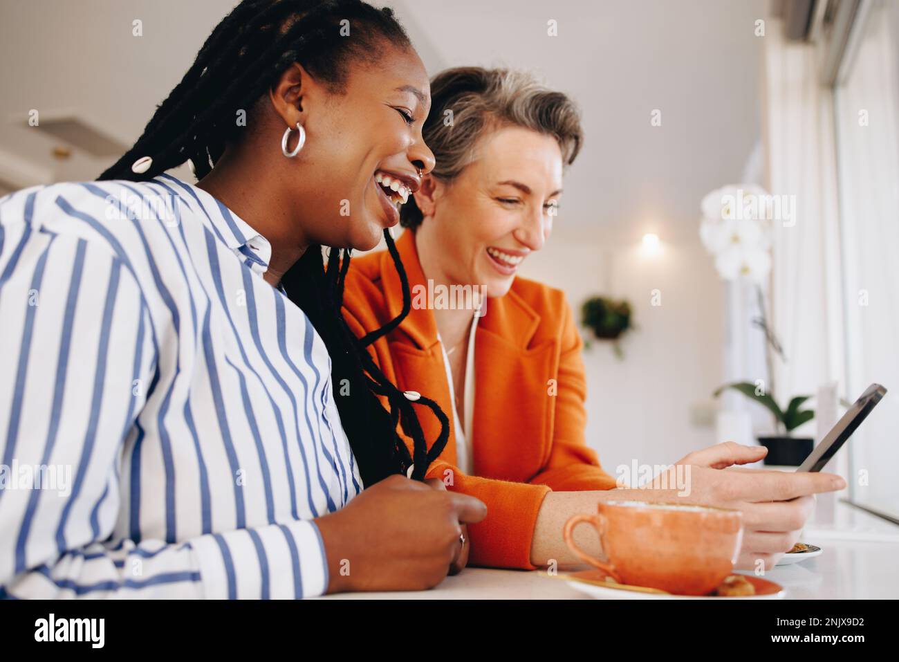 Zwei weibliche Kollegen, die fröhlich lächeln, während sie ein Smartphone in einem Bürocafé gemeinsam nutzen. Glückliche Geschäftsfrauen, die ihre Kaffeepause zusammen genießen Stockfoto