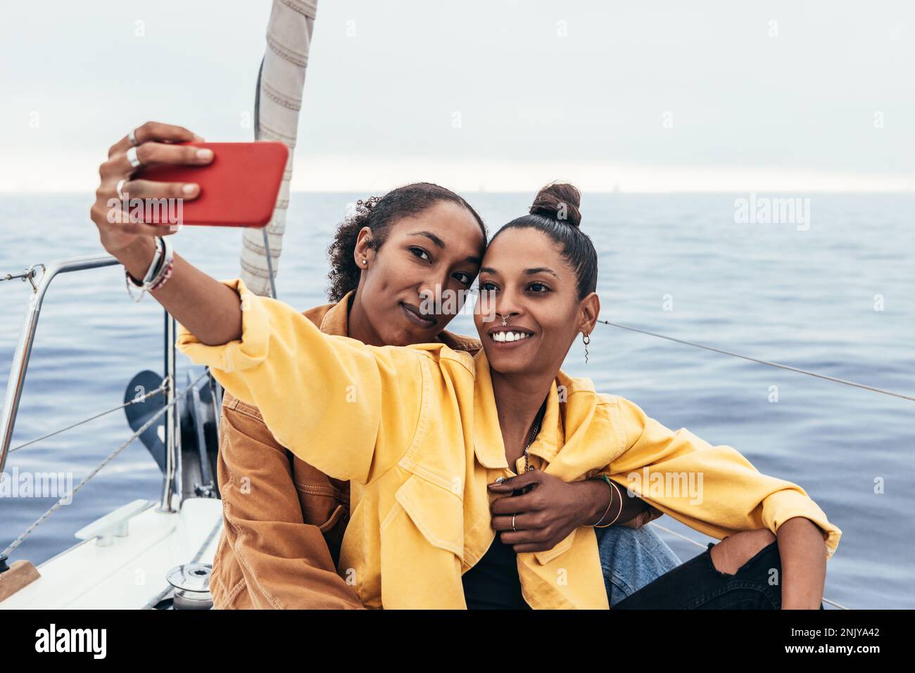 Positive afroamerikanische Freundinnen in lässigen Outfits, die sich umarmen und Selfie auf dem Smartphone machen, während sie sich zusammen auf der Yacht ausruhen Stockfoto