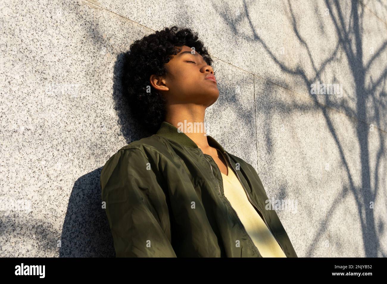 Von unten selbstbewusster junger asiatischer Mann mit Afro-Frisur in gelbem Sweatshirt mit Augen geschlossen gegen geflieste Wand mit Schatten im Sonnenlicht Stockfoto