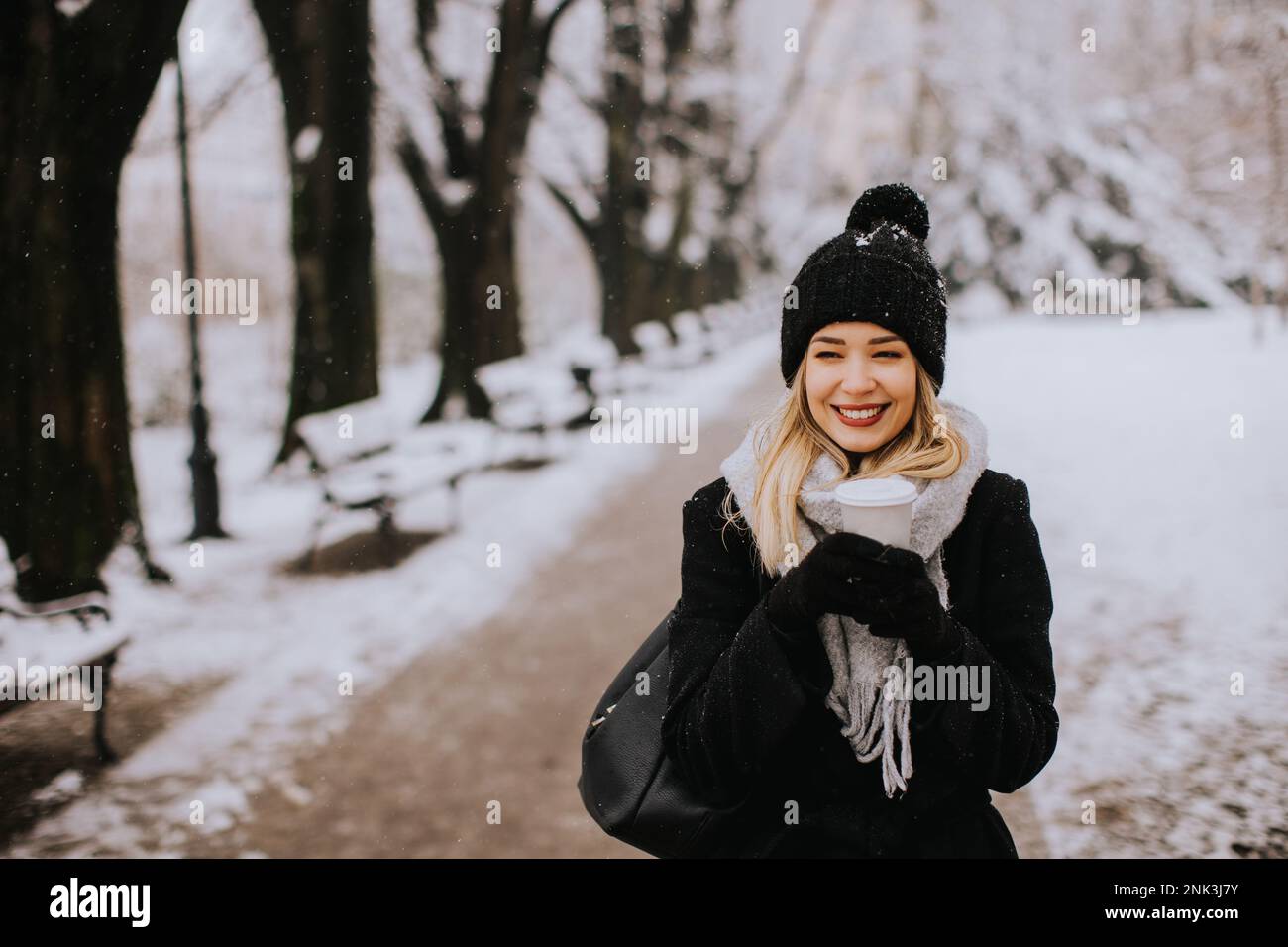 Eine junge Frau steht in einem Winterwunderland, trägt warme Kleidung und trinkt heißen Kaffee zum Mitnehmen Stockfoto