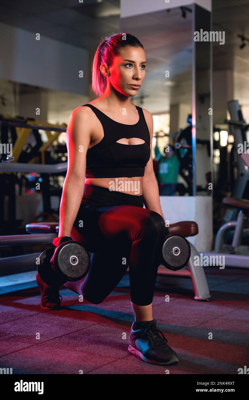 Eine 30s-jährige Frau mit schwarzen Oberteilen und Strumpfhosen, die in einem Fitnessstudio Ausfallschritte mit Kurzhanteln macht. Stockfoto