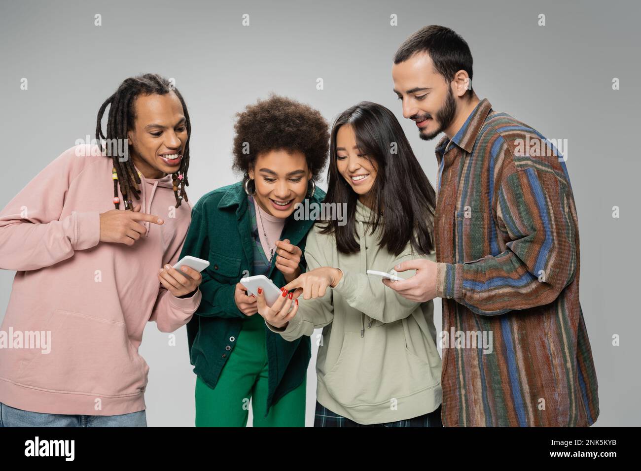 Fröhliche asiatische Frau zeigt auf Smartphone in der Nähe trendiger multiethnischer Freunde isoliert auf grauem Stockbild Stockfoto