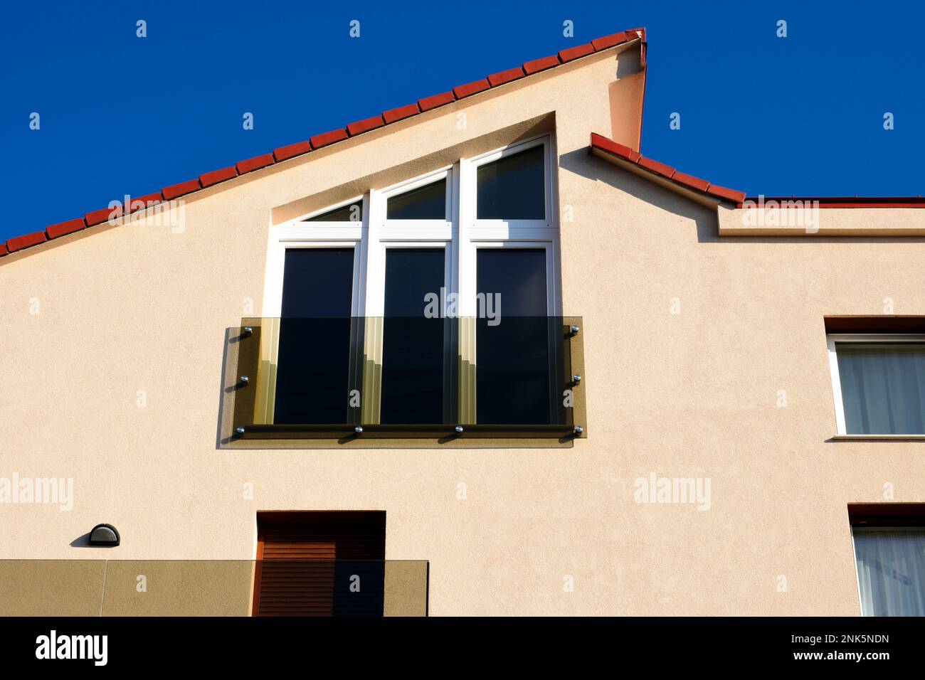 Glasbalustrade auf einem kleinen französischen Balkon mit großen trapezförmigen Fenstern. Stuckfassade. Abgeschrägtes Tondach. Modernes Wohnarchitekturkonzept Stockfoto