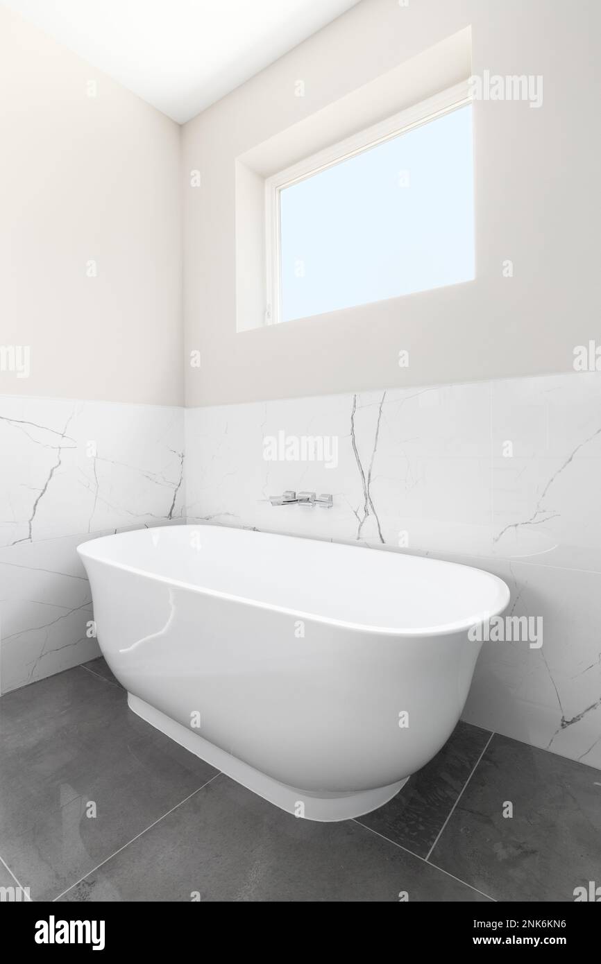 Eine freistehende Badewanne mit eingebautem Wasserhahn auf einem dunkelgrauen Fliesenboden, weiße Marmorwände und ein Fenster darüber. Stockfoto
