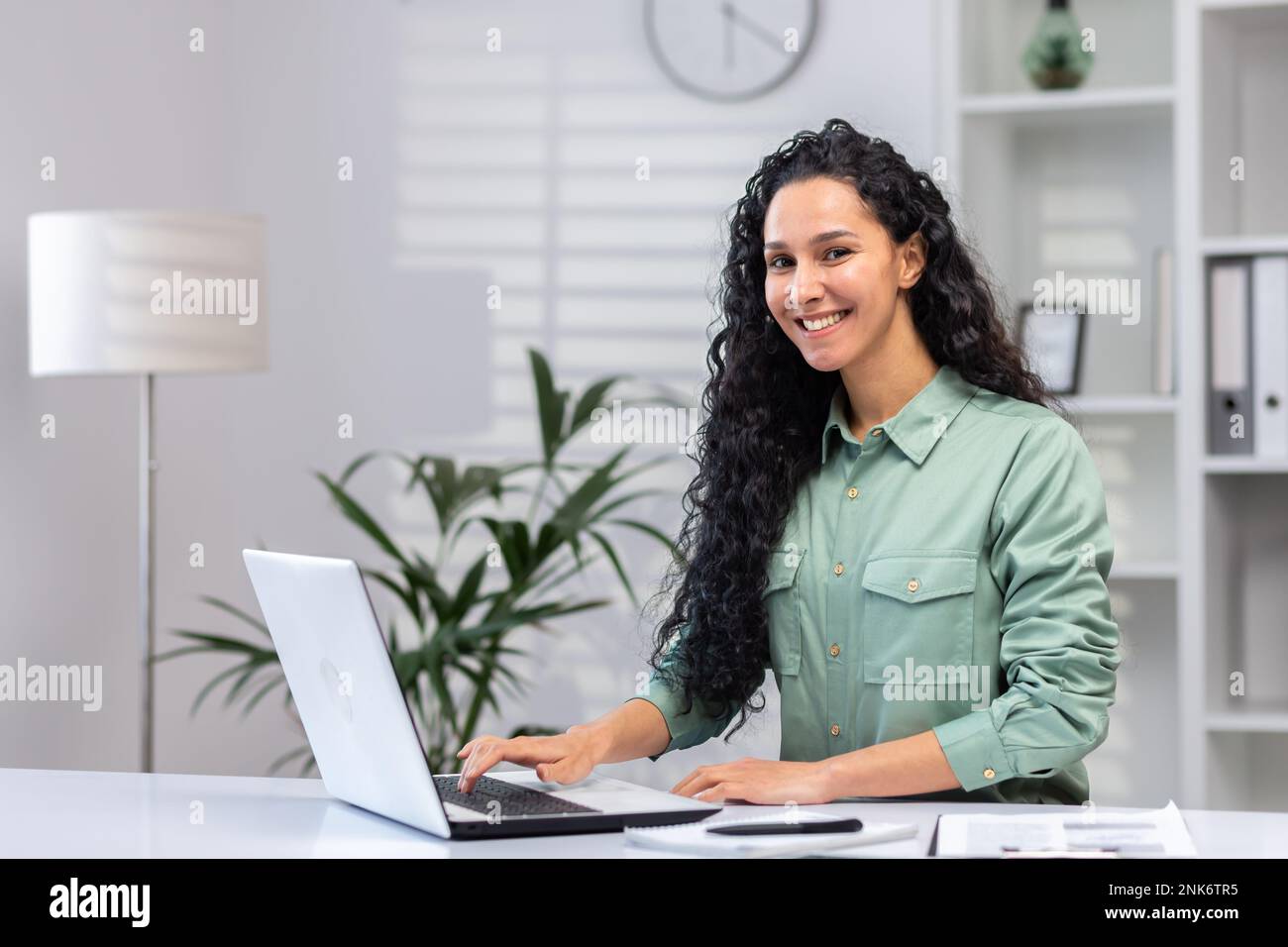 Porträt einer erfolgreichen, schönen Geschäftsfrau, einer hispanischen Frau im Heimbüro, die lächelt und in die Kamera schaut, einer Frau bei der Arbeit, die mit einem Laptop über die Tastatur tippt. Stockfoto