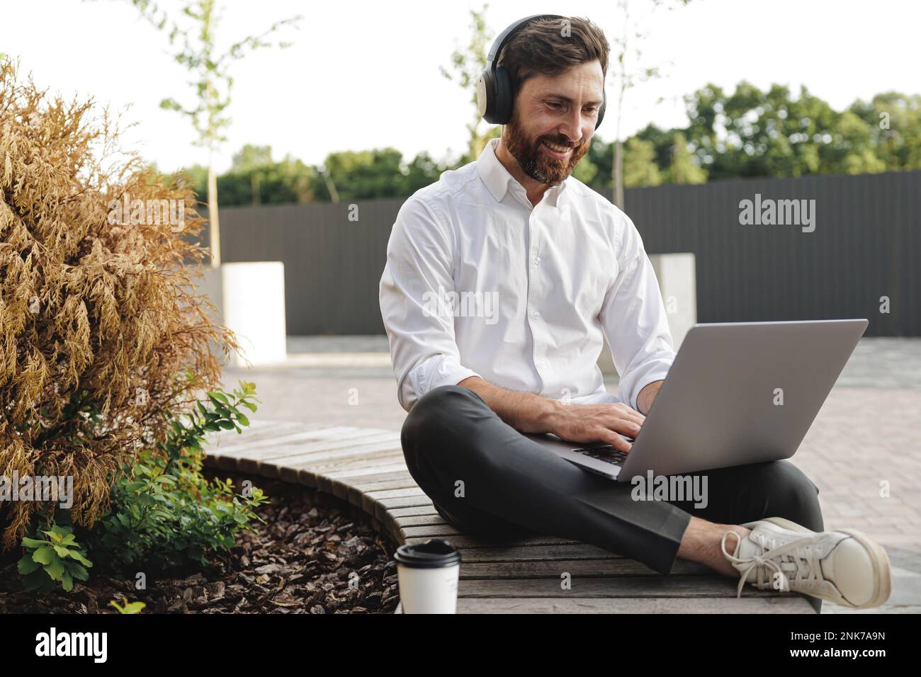Eleganter Unternehmer mit Kopfhörern, der auf einer Bank in der Stadt sitzt und E-Mails auf dem Laptop tippt. Glücklicher Mann, der aufmerksam auf den Bildschirm schaut und arbeitet Stockfoto