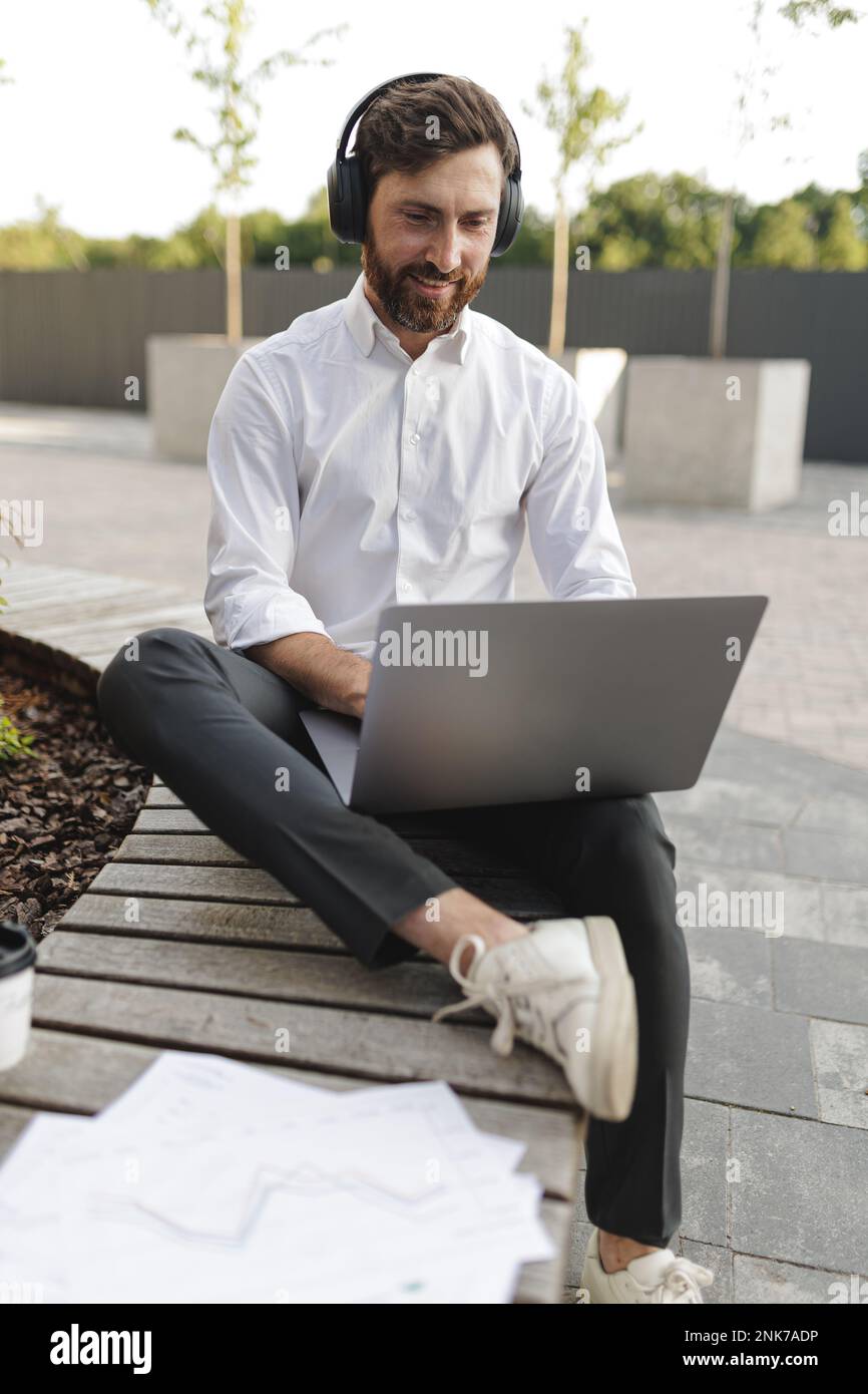 Positiver Büroangestellter mit Kopfhörern, der mit freudigem Gesichtsausdruck auf den Laptop-Bildschirm schaut. Eleganter Mann, der am Computer arbeitet, während er auf Holz sitzt Stockfoto