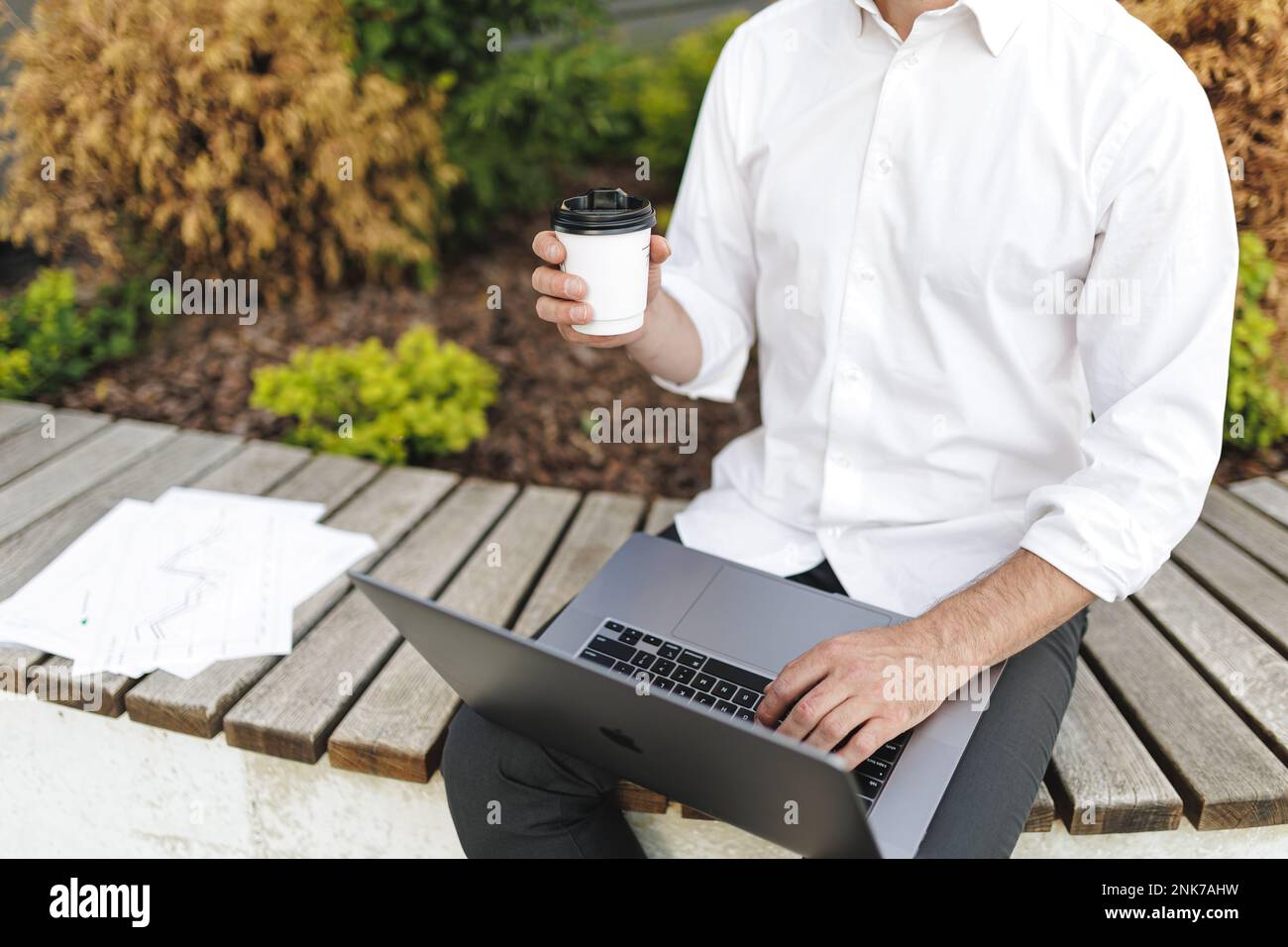 Männlicher Büroangestellter, der ein tragbares Notebook verwendet, um Informationen zu suchen, während er Kaffee zum Mitnehmen genießt. Zugeschnittener Blick auf den Mann in formeller Kleidung, der im Freien arbeitet Stockfoto