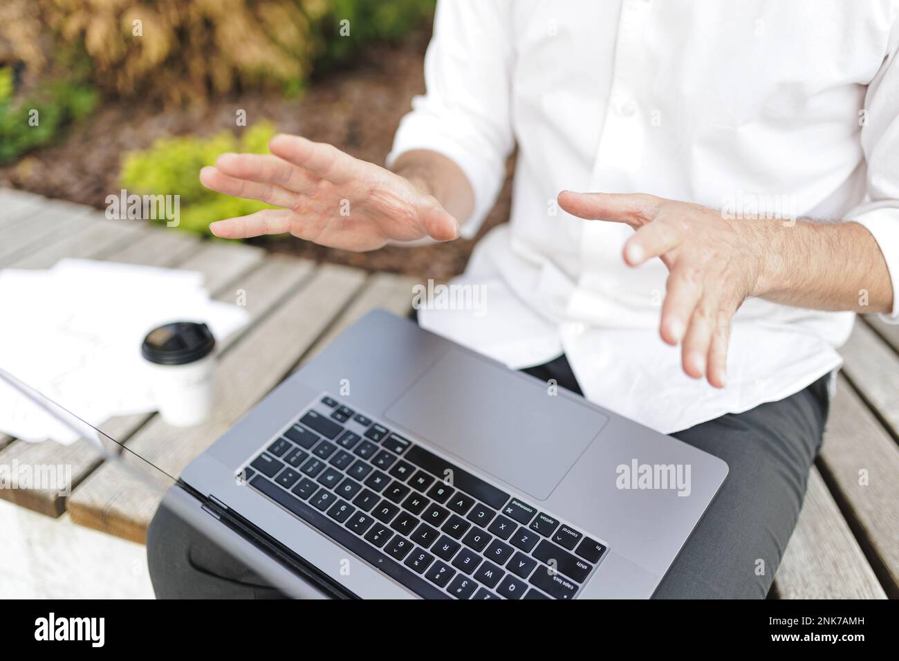 Nahaufnahme eines Mannes in formeller Kleidung bei einer Online-Videokonferenz auf einem tragbaren Laptop. Männlich sitzt draußen auf einer Holzbank und hat aktiv Gesturin Stockfoto
