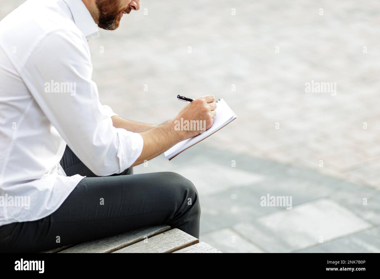 Kurzer Blick auf einen bärtigen Geschäftsmann, der auf einer Bank in der Stadt sitzt und sich Notizen macht. Stylischer Mann in formeller Kleidung, der kreative Ideen aufschreibt Stockfoto