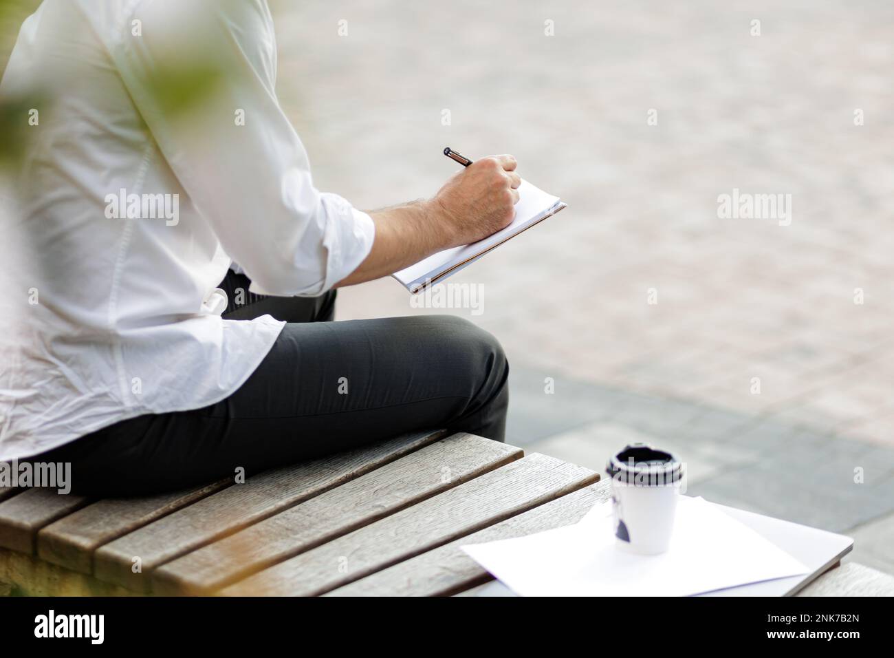 Stilvoller Mann, der einen neuen Geschäftsplan in ein Tagebuch schreibt. Büroangestellter in formeller Kleidung sitzt auf einer Bank neben Laptop, Kaffeetasse und Dokumenten während der Arbeit in Stockfoto