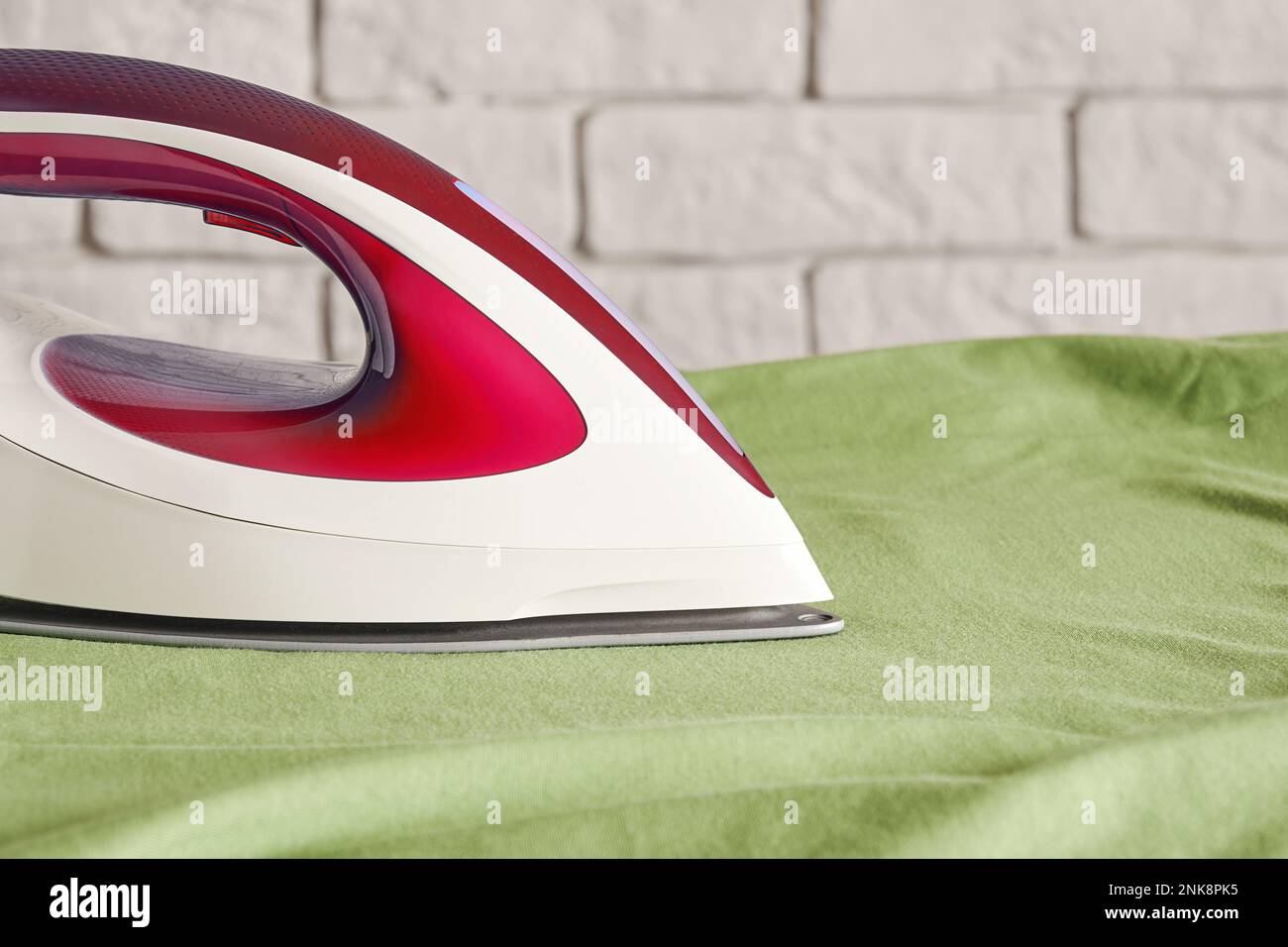 Modernes rotes Bügeleisen auf dem Bügelbrett, grünes Hemd - Nahaufnahme. Haushaltsgeräte. Platz für Text kopieren Stockfoto