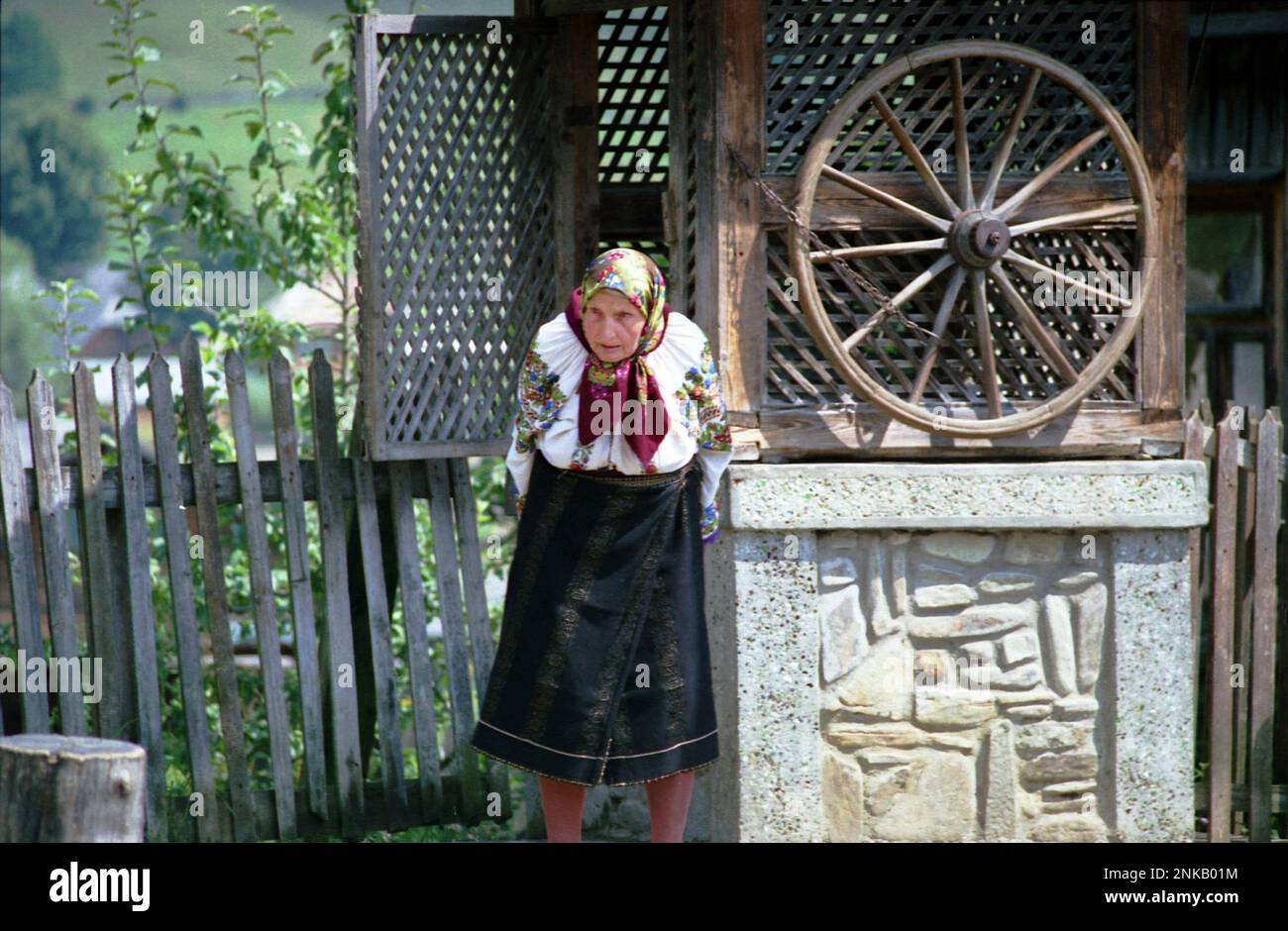 Straja, Suceava County, Rumänien, 1998. Ältere Frau in traditionellem Kostüm in der Nähe eines Brunnens mit Rad. Stockfoto