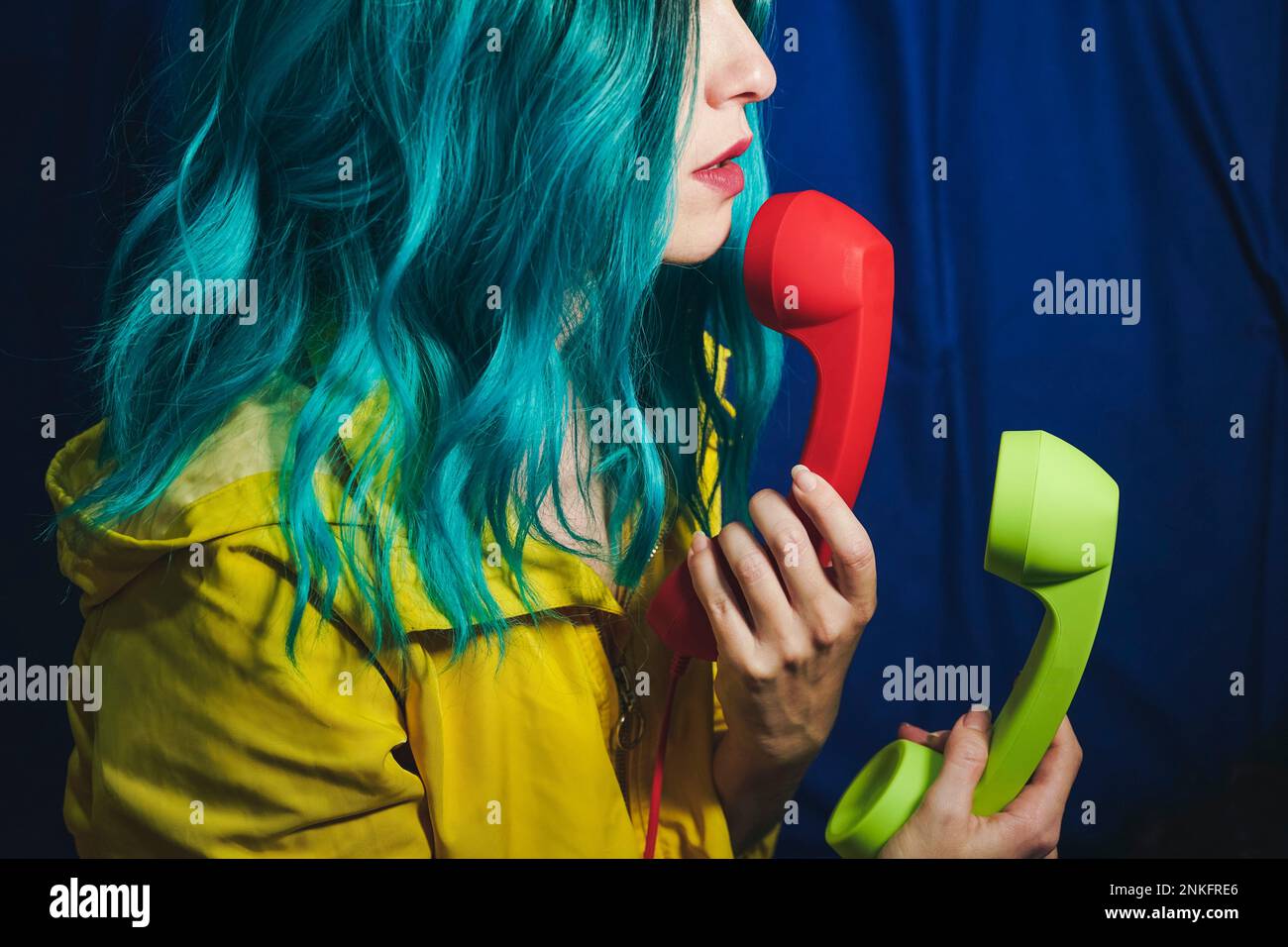 Frau mit gefärbtem Haar, die rote und grüne Telefonhörer vor blauem Hintergrund hält Stockfoto