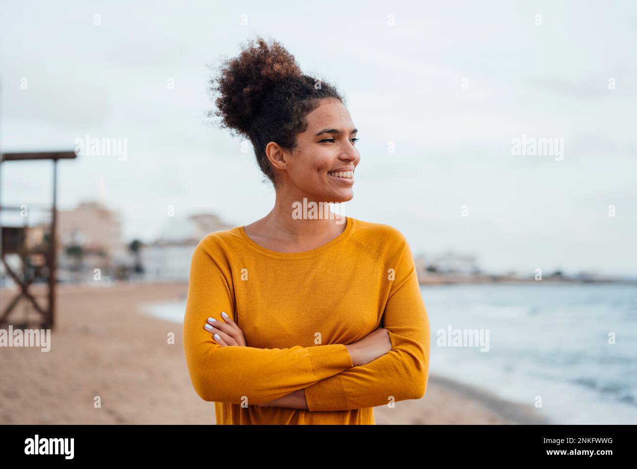 Lächelnde junge Frau, die am Strand mit gekreuzten Armen stand Stockfoto