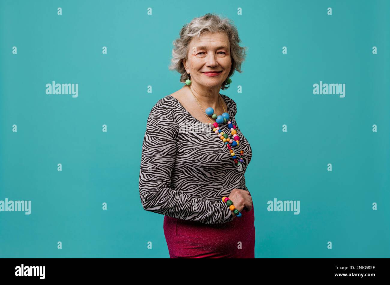 Lächelnde Seniorin mit mehrfarbigem Schmuck vor türkisfarbenem Hintergrund Stockfoto