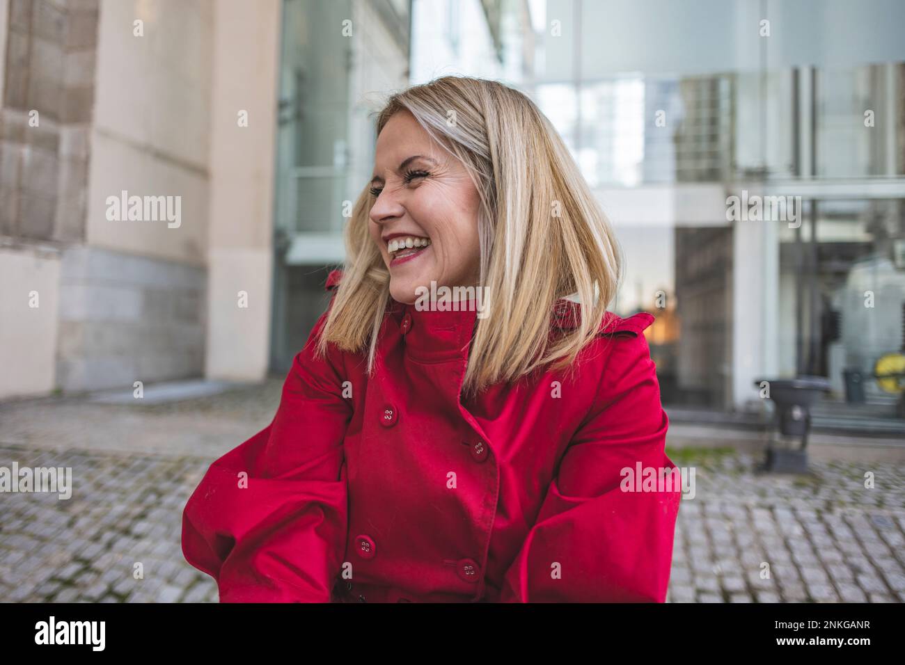 Reife Frau mit rotem Mantel, die vor dem Gebäude lacht Stockfoto