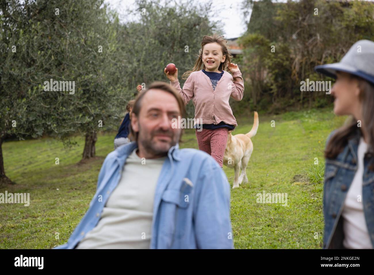 Mann und Frau sitzen auf dem Gras, und die Tochter läuft im Hintergrund Stockfoto