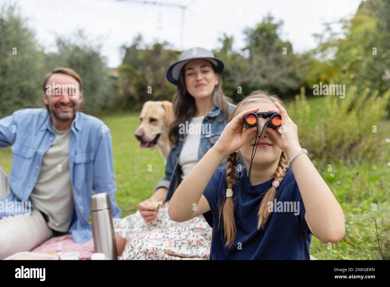 Ein Mädchen, das durch ein Fernglas schaut, während Eltern im Hintergrund beim Familien-Picknick sitzen Stockfoto