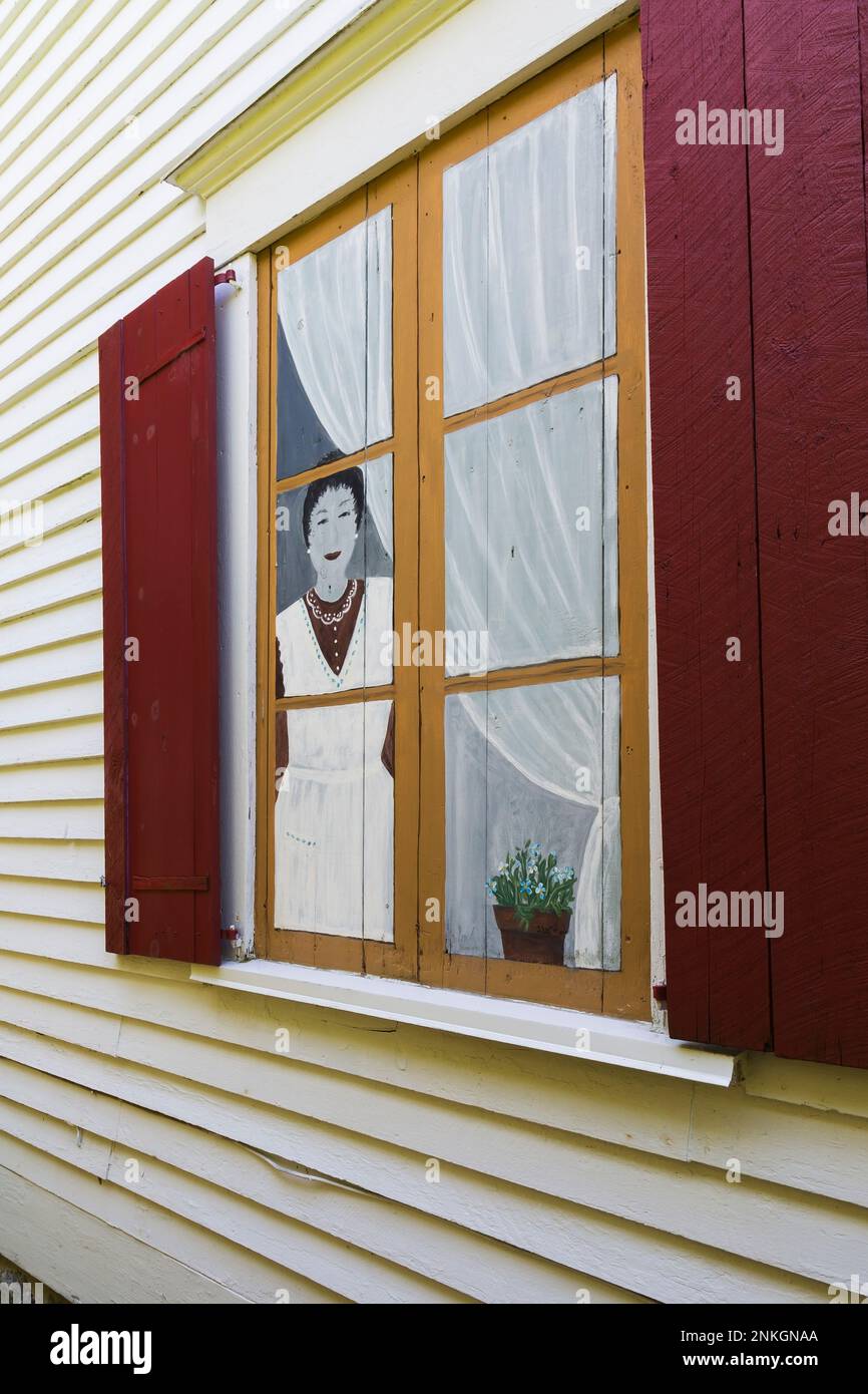 Frau steht in einer gefälschten bemalten Fensterszene mit roten hölzernen Fensterläden an der äußeren Seitenwand des alten, um 1840 Jahre alten, weißen horizontalen Holzplanken-Hauses. Stockfoto