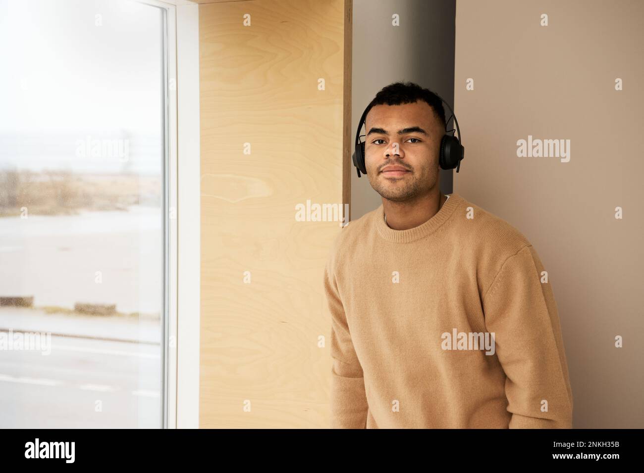 Lächelnder junger Mann mit Kopfhörern, der am Fenster steht Stockfoto