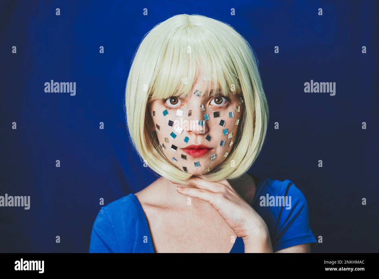 Blonde Frau mit kleinen Spiegeln im Gesicht vor blauem Hintergrund Stockfoto