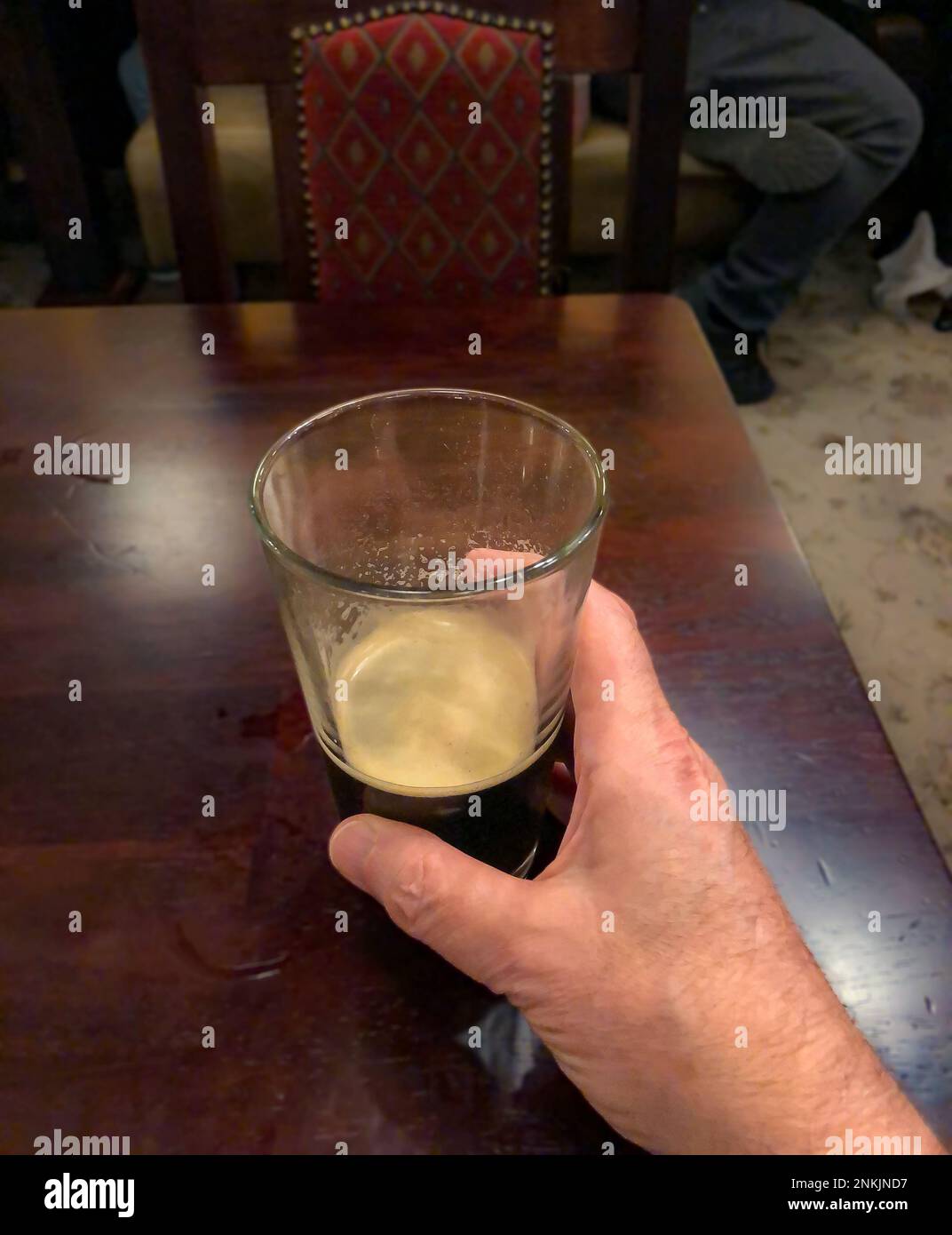 Nahaufnahme eines fast leeren Glases Bier mit Schaumschicht und menschlicher Hand auf einem Holztisch in einem Restaurant. Stockfoto