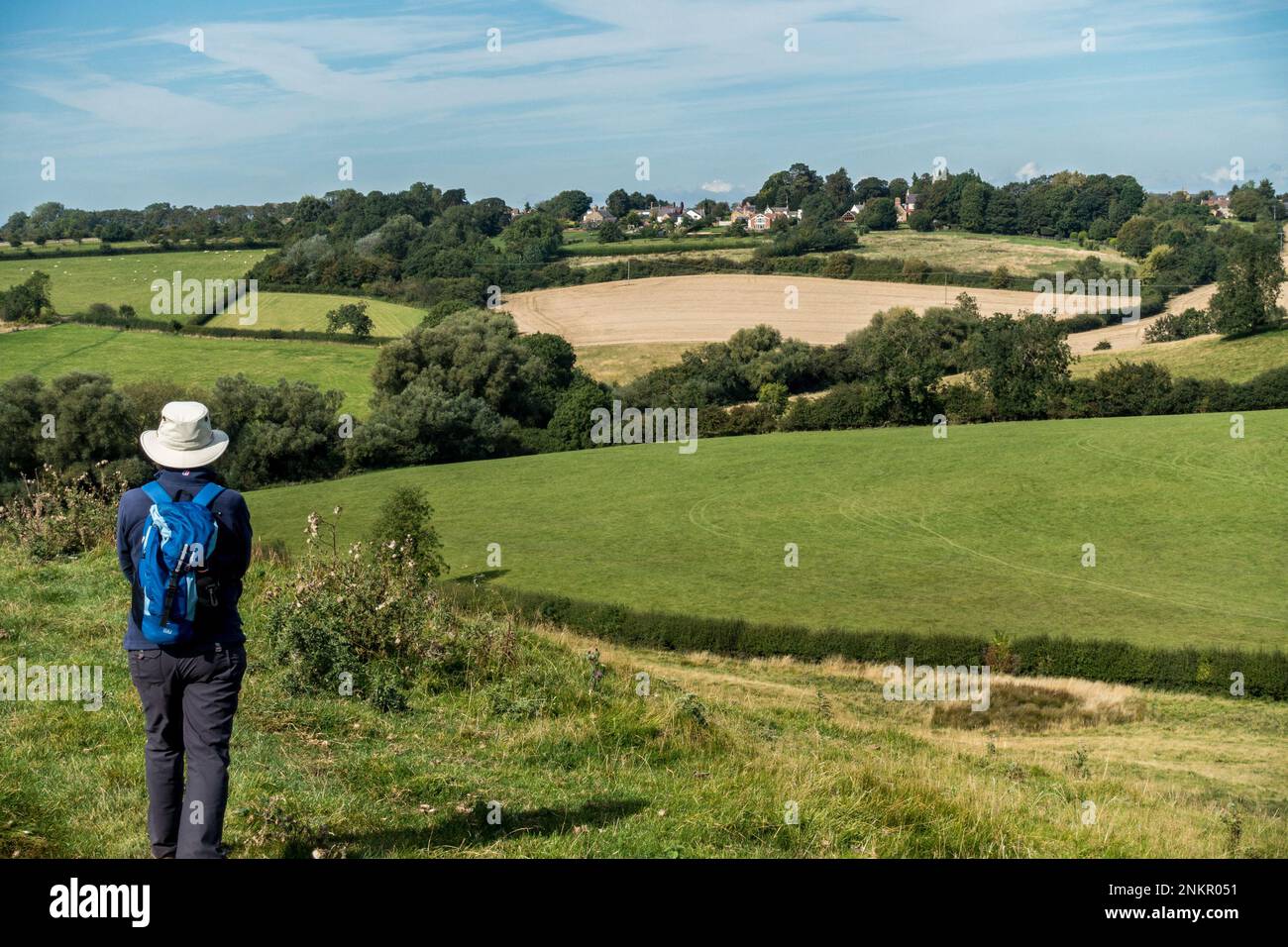 Rambler geht in Richtung des auf einem Hügel gelegenen Dorfes Burrough on the Hill, das aus der Ferne über Leicestershire Farm Fields, England, Großbritannien, zu sehen ist Stockfoto