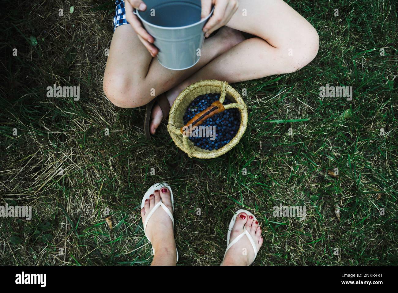 Blick von oben auf ein Mädchen, das mit überkreuzten Beinen auf einem Graseimer sitzt, während Osterkorb und Mutter in der Nähe stehen Stockfoto
