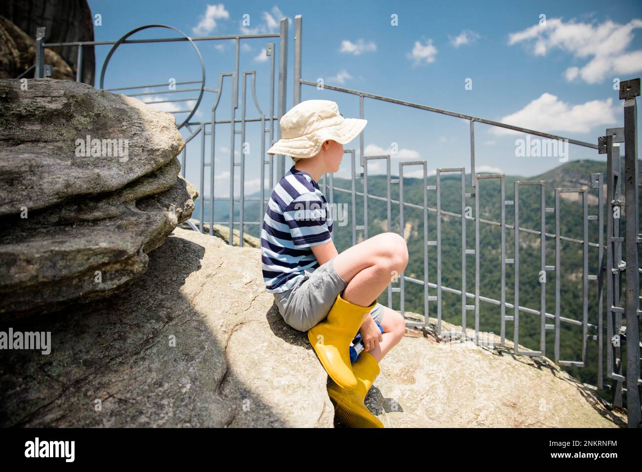 Ein junger Junge mit Hut und gummistiefeln, der auf einem Felsen sitzt und die Aussicht im Urlaub betrachtet Stockfoto