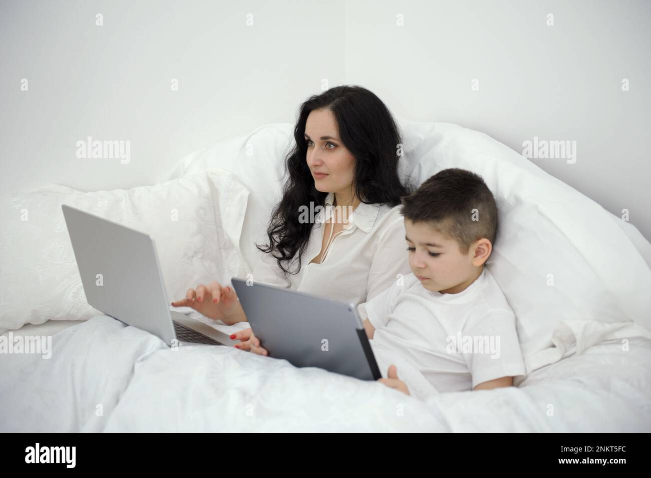 Work Home Online Frau unter Decke Arbeit auf Laptop neben Sohn Spiele auf Tablet zu Hause krank zu Hause Get Family Isolation Quarantäne weiß Bettdecke schwarze Haare mutter und Sohn Stockfoto