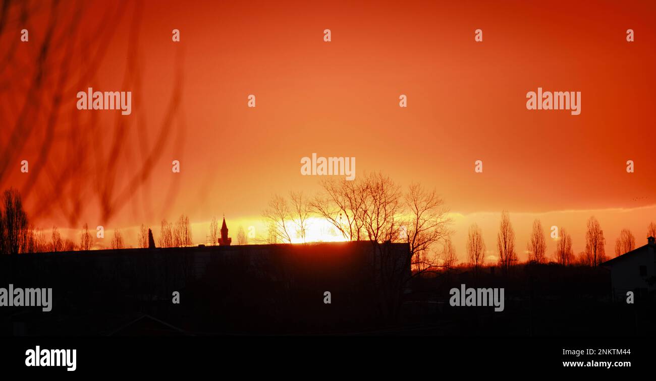 Sonnenaufgang und suggestives Licht bei bewölktem Himmel mit Nuancen von Orange bis Blau und Hinterleuchtsilhouette aus Holz und Gebäude Stockfoto