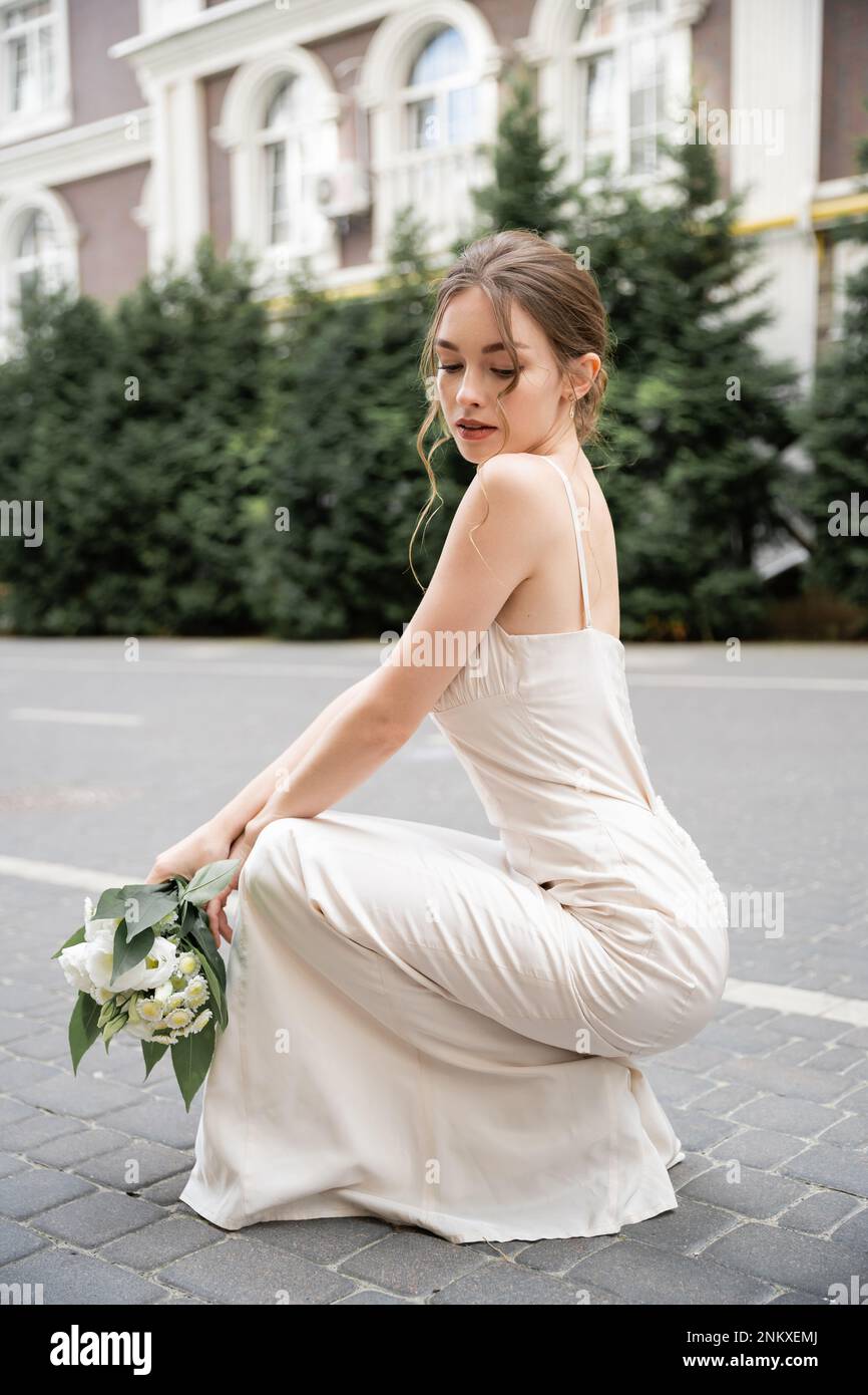 Junge Braut im Hochzeitskleid hält einen Blumenstrauß mit Blumen, während sie draußen sitzt, Stockbild Stockfoto