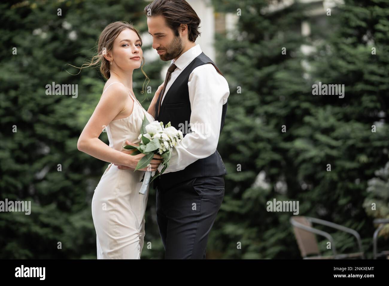 Bärtiger Bräutigam in Weste umarmt Braut in Hochzeitskleid mit Blumenstrauß, Stockbild Stockfoto