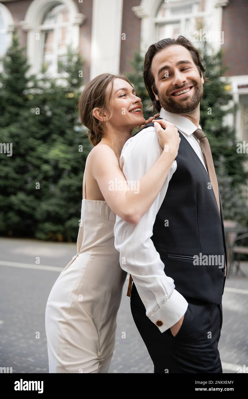 Fröhliche junge Frau im Hochzeitskleid, die einen glücklichen Bräutigam in Weste umarmt, Stockbild Stockfoto
