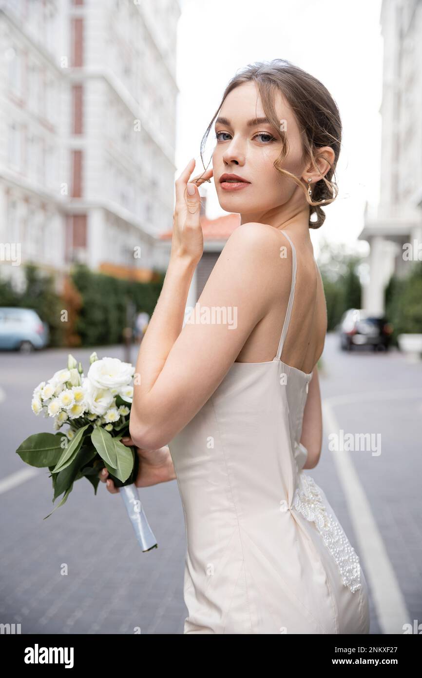 Junge Frau in weißem Kleid hält Hochzeitsstrauß und schaut in die Kamera, Stockbild Stockfoto