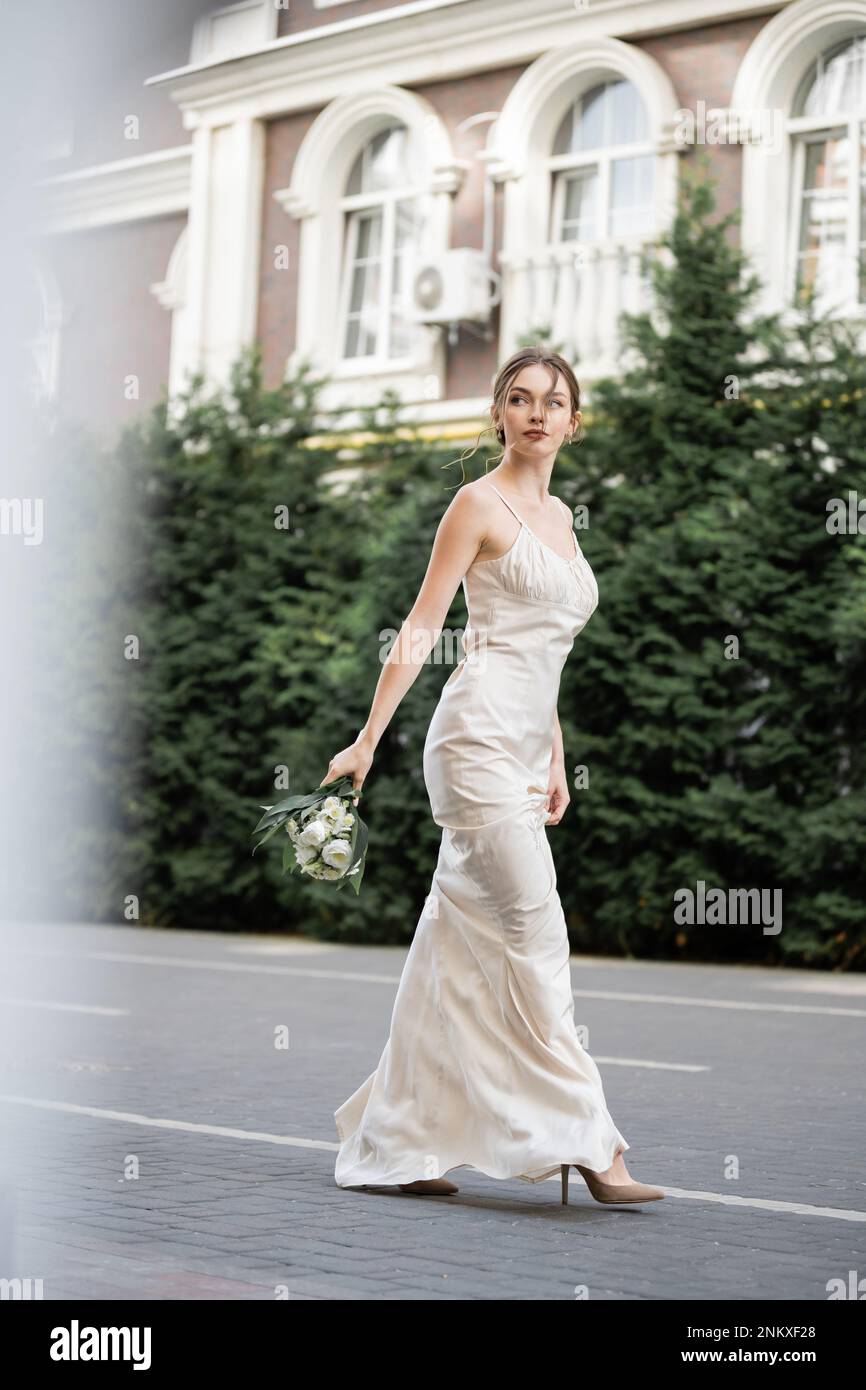 Lange junge Braut in weißem Kleid, die einen Hochzeitsstrauß voller blühender Blumen hält und nach draußen geht, Stockbild Stockfoto