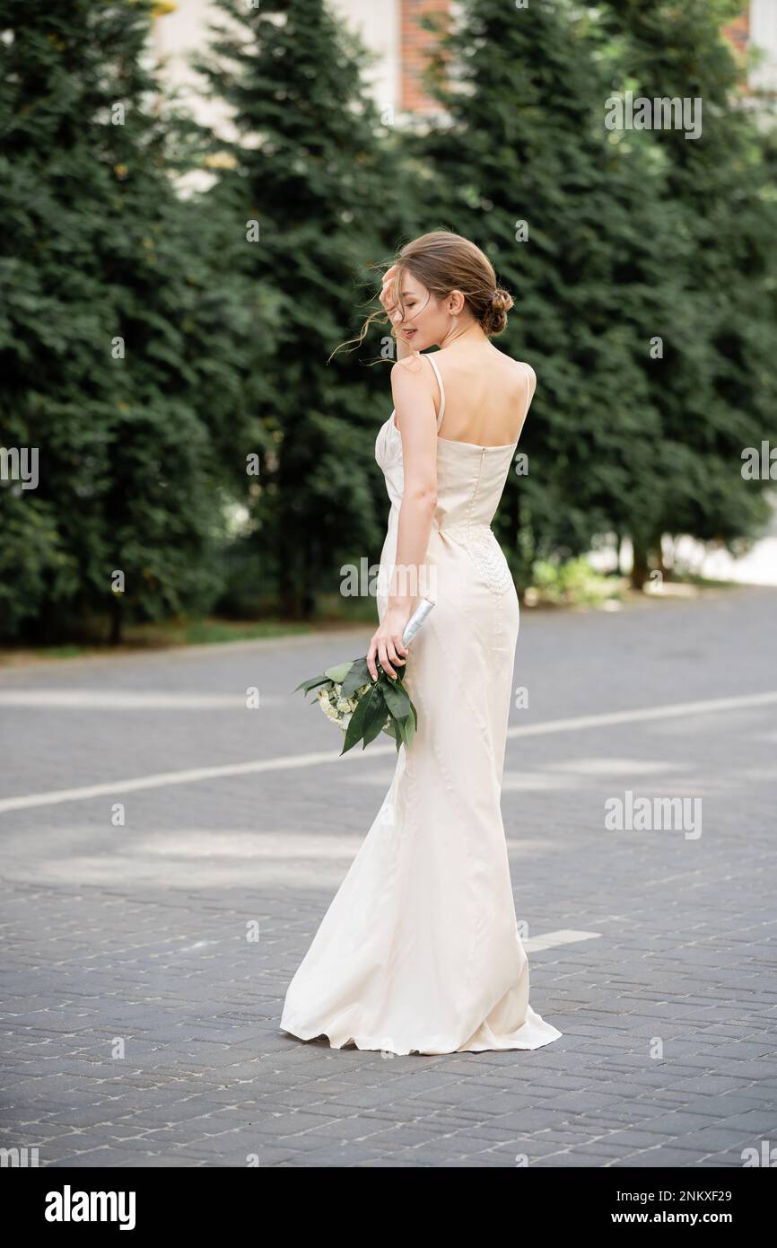 Die ganze Länge der glücklichen Braut in weißem Kleid hält einen Hochzeitsstrauß und steht auf der Straße, Bild der Aktie Stockfoto