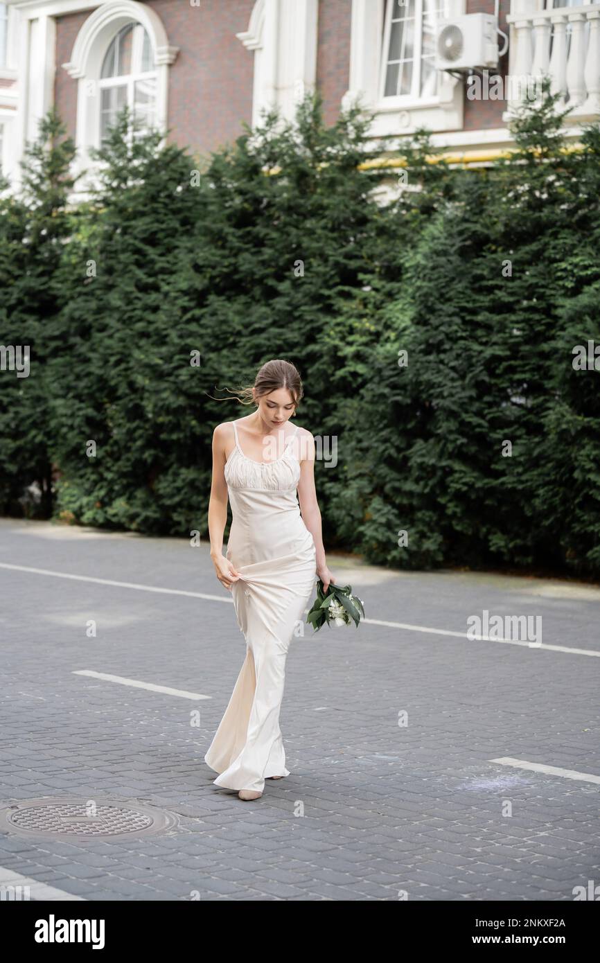 Lange junge Braut in weißem Kleid mit Blumenstrauß und Spaziergängen auf der Straße, Bild der Börse Stockfoto