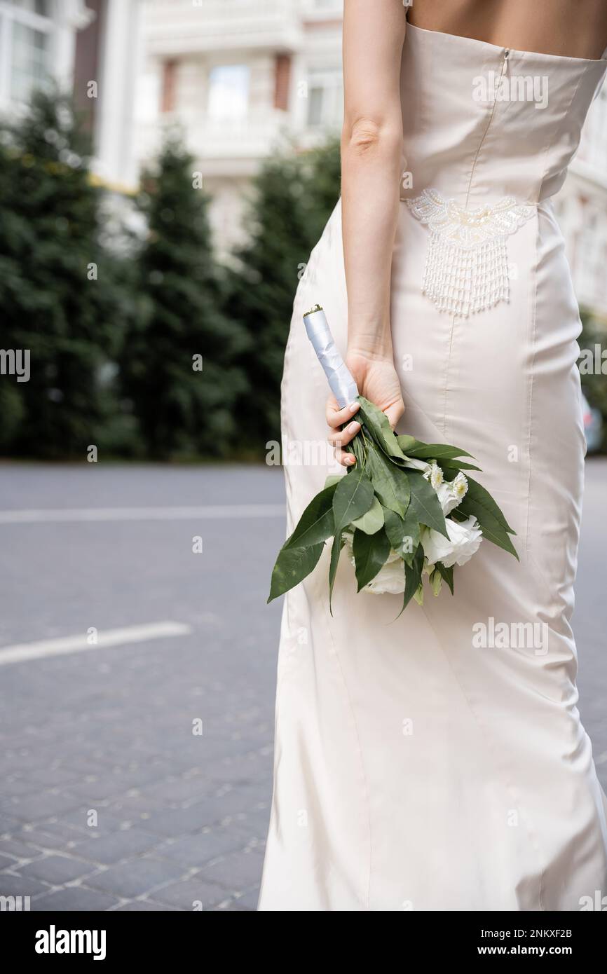 Zugeschnittener Blick auf eine junge Frau in weißem Kleid mit Hochzeitsstrauß hinter dem Rücken, Stockbild Stockfoto